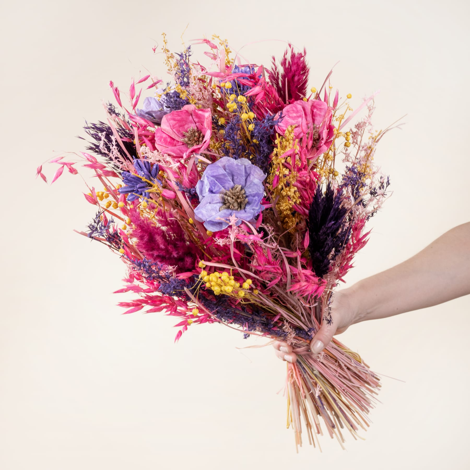 Ein lebendiger Strauß bunter Trockenblumen, darunter rosa, violette und gelbe Blüten, in der Hand gehalten vor einem schlichten, hellen Hintergrund.