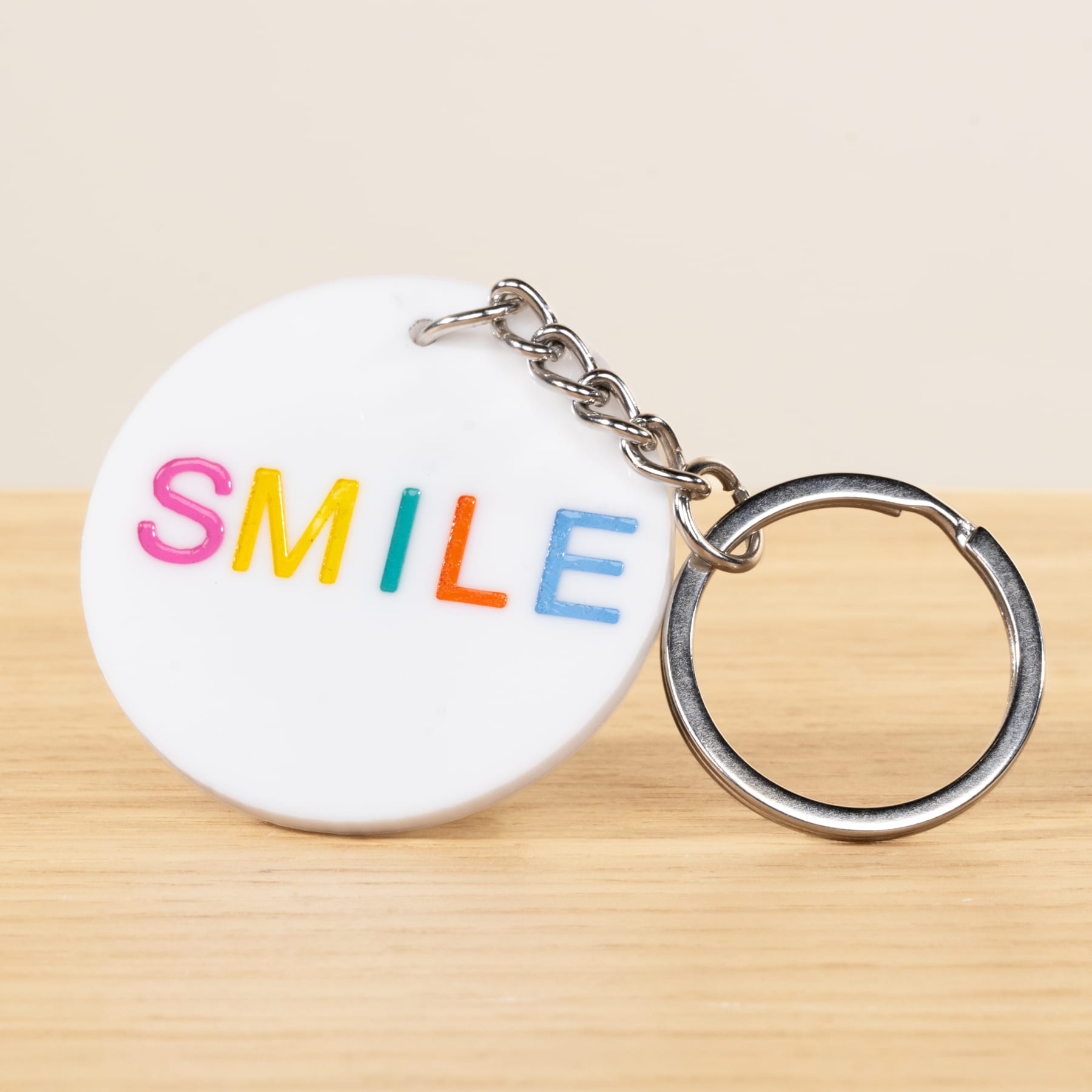 Auf einer Holzoberfläche liegt ein weißer, runder Schlüsselanhänger mit dem Wort „SMILE“ in bunten Buchstaben.