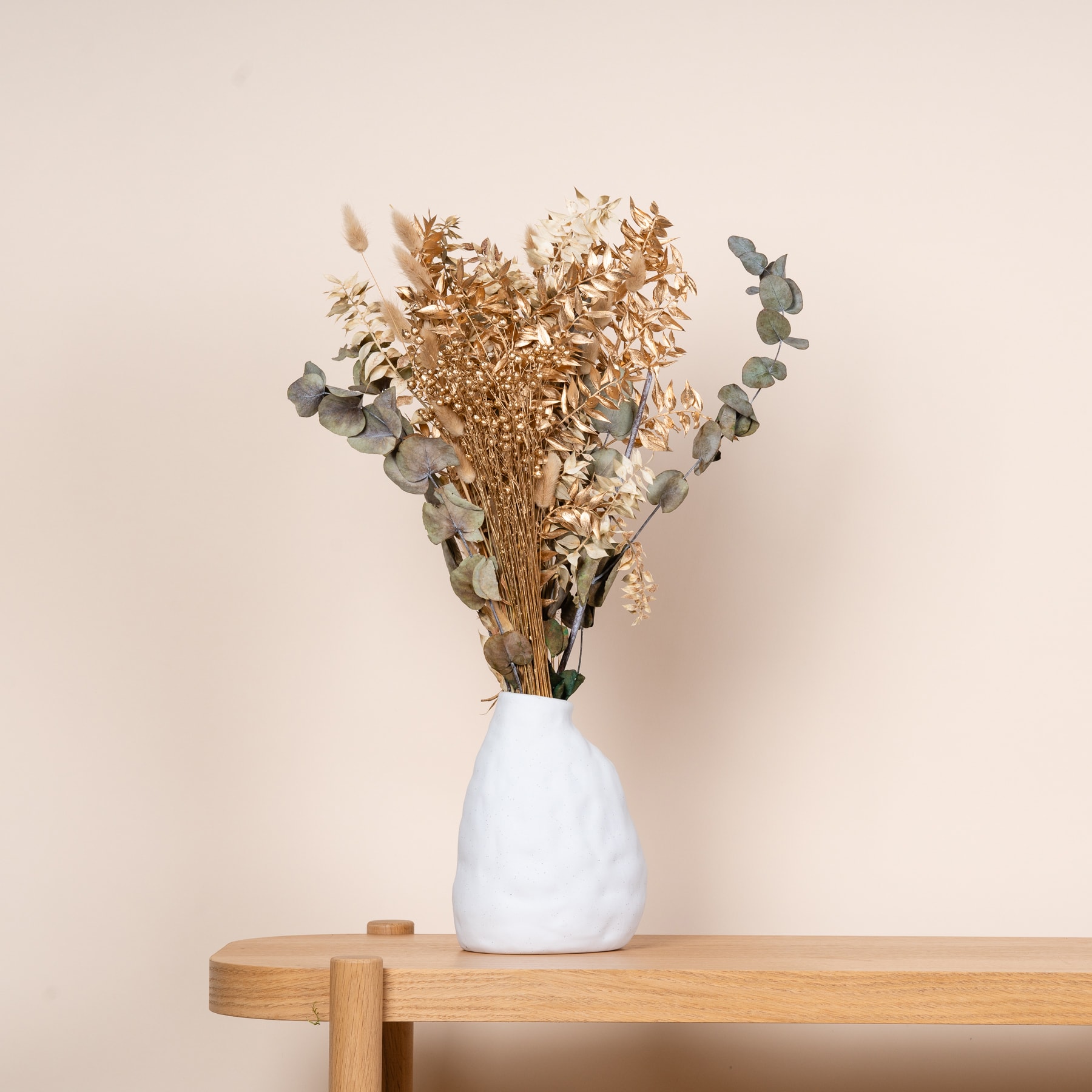 Eine Vase mit einem Strauß getrockneter Blumen steht auf einem Holztisch vor einem neutralen beigen Hintergrund.