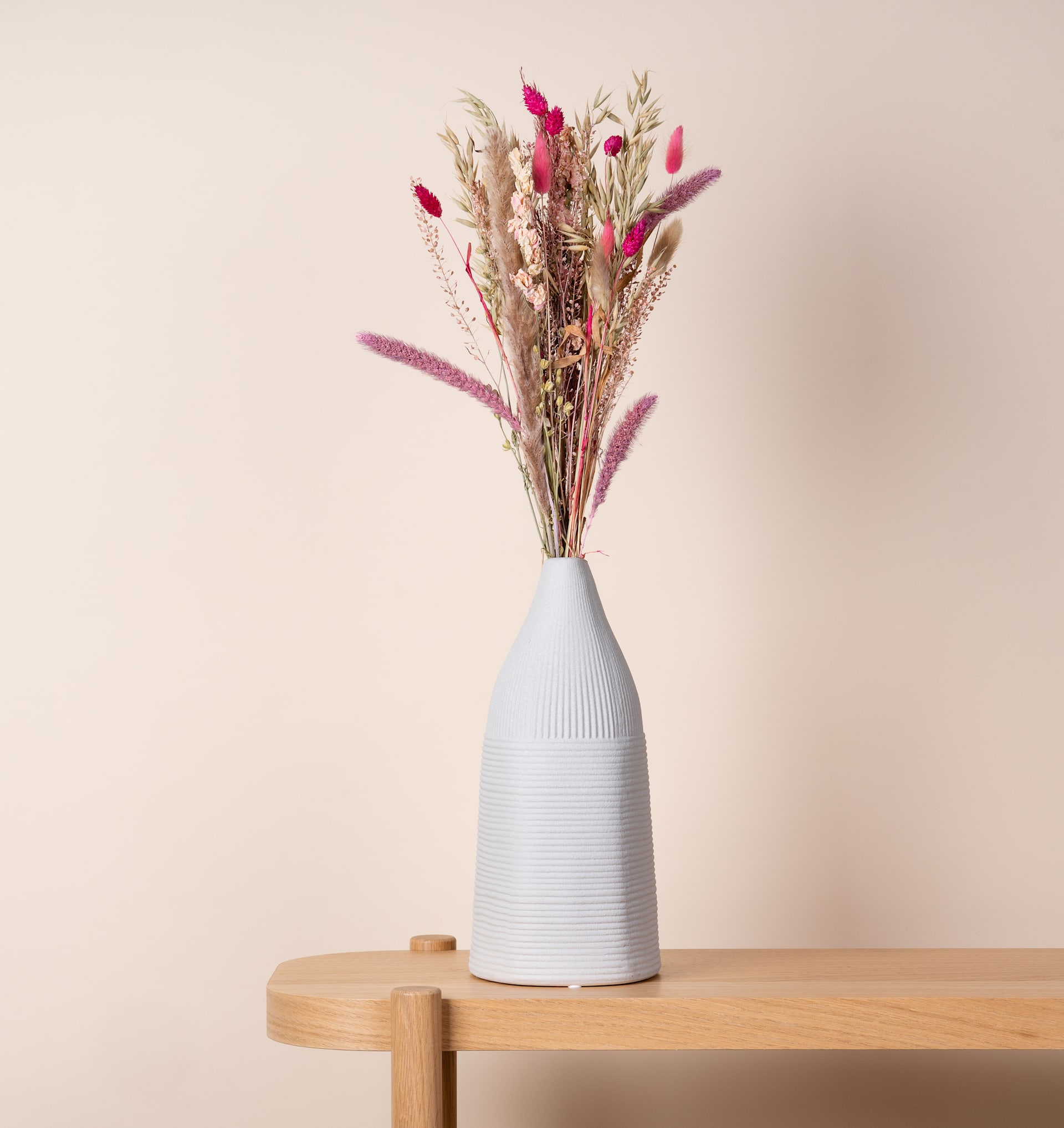 Eine weiße Keramikvase mit einem farbenfrohen Strauß getrockneter Blumen und Pflanzen steht auf einem Holztisch vor einem neutralen beigen Hintergrund.