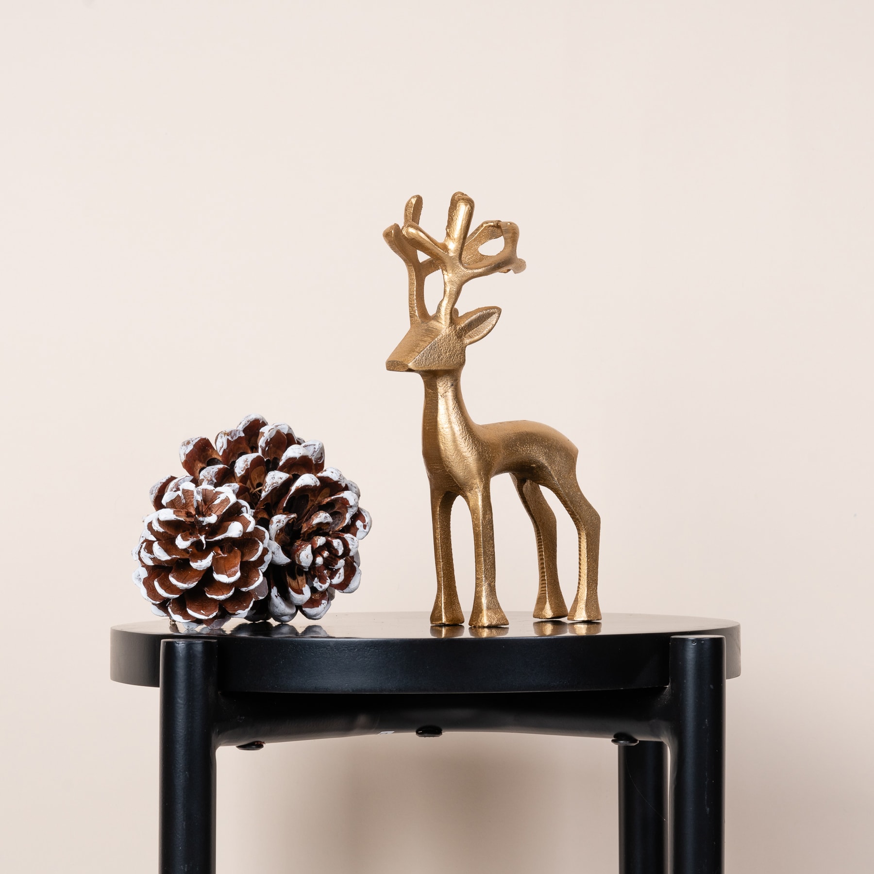 Eine goldene Hirschfigur und ein Tannenzapfen stehen auf einem schwarzen Holztisch vor einem schlichten beigen Hintergrund.