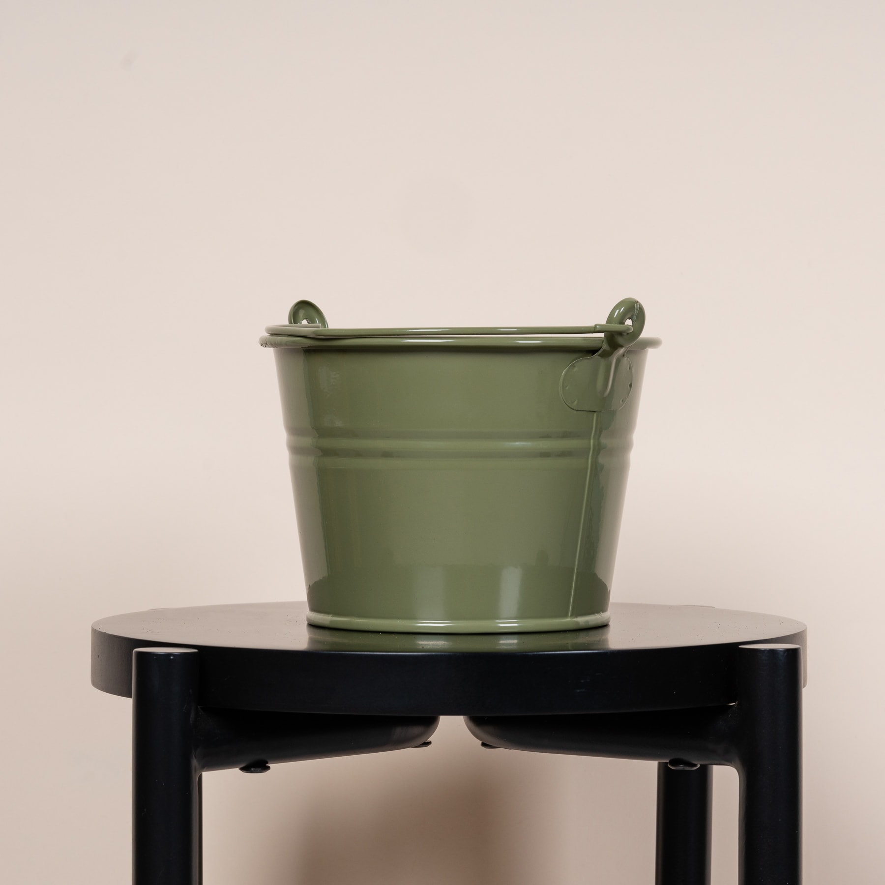 Ein grüner Metalleimer steht auf einem schwarzen Holzständer vor einem schlichten beigen Hintergrund.
