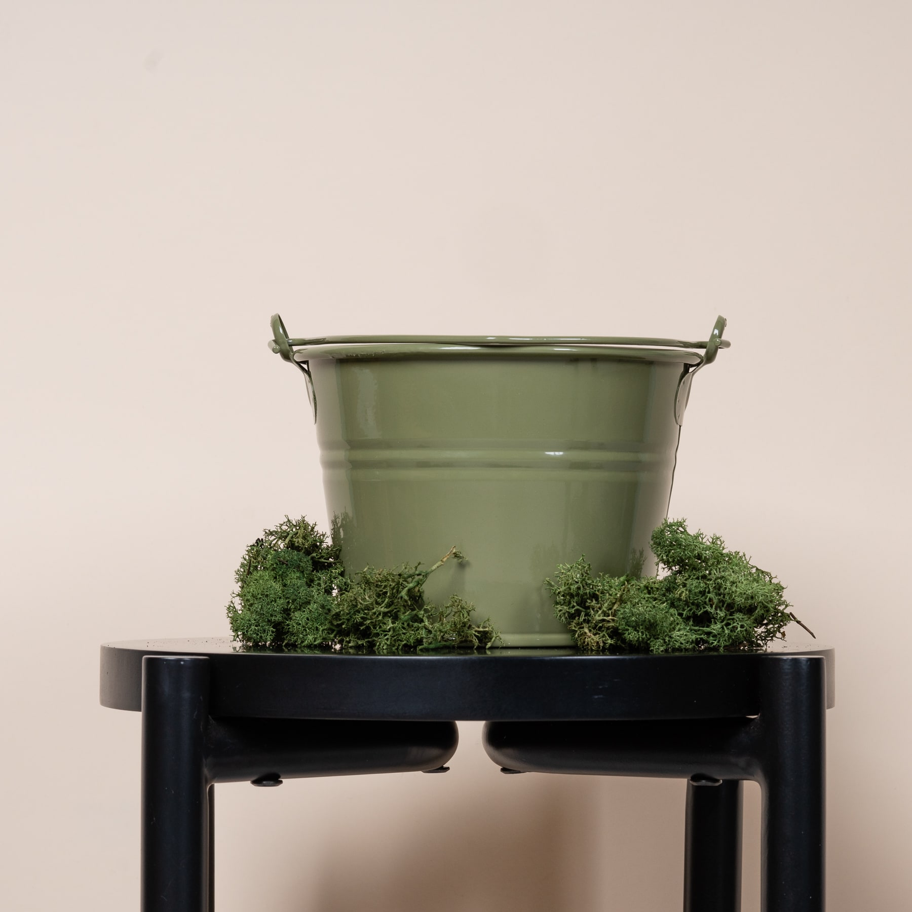 Ein grüner Metalleimer voller üppiger grüner Pflanzen steht auf einem schwarzen Metallständer vor einem schlichten beigen Hintergrund.