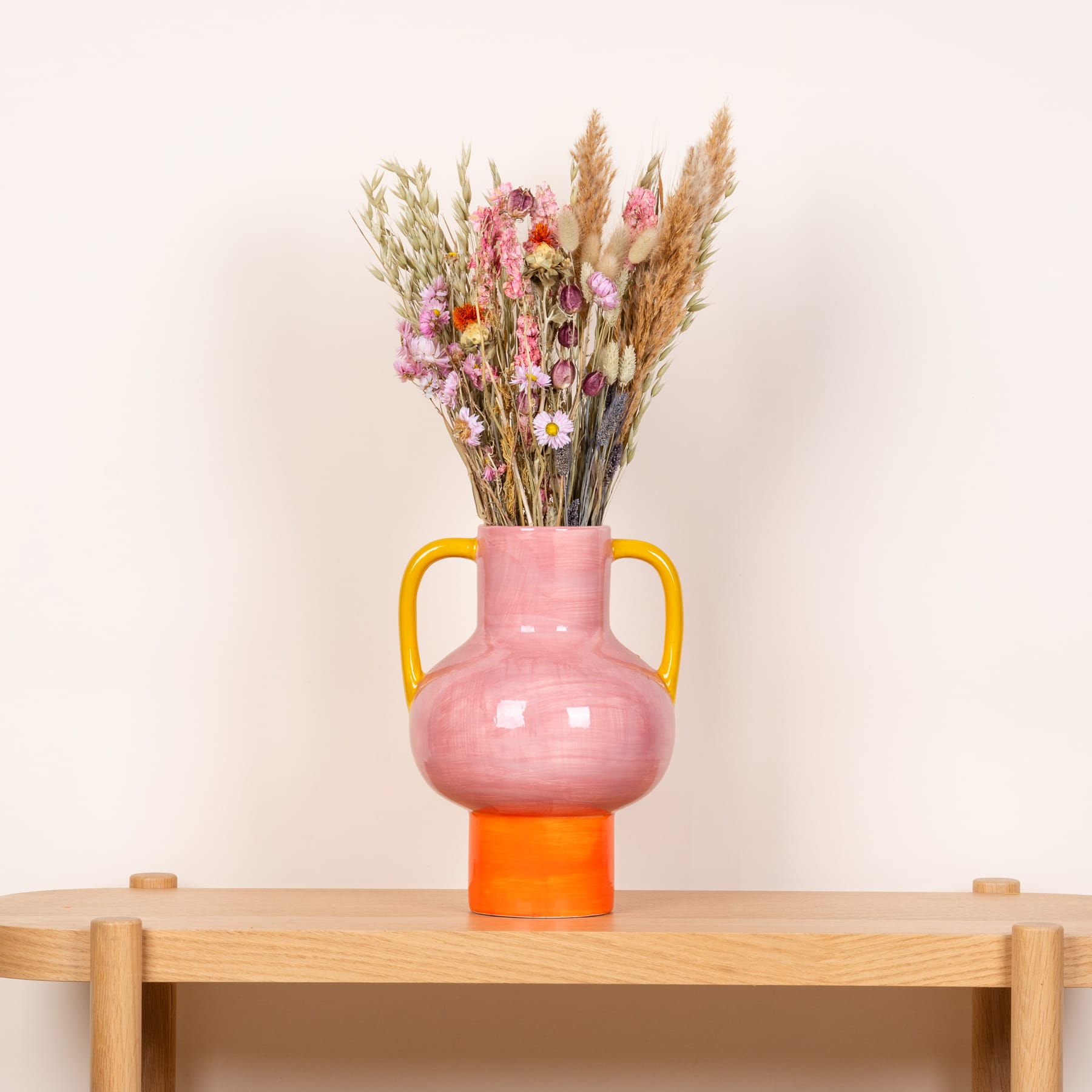 Eine rosa Keramikvase mit einem Strauß getrockneter Blumen und Gräser steht auf einem Holzregal vor einem schlichten beigen Hintergrund.