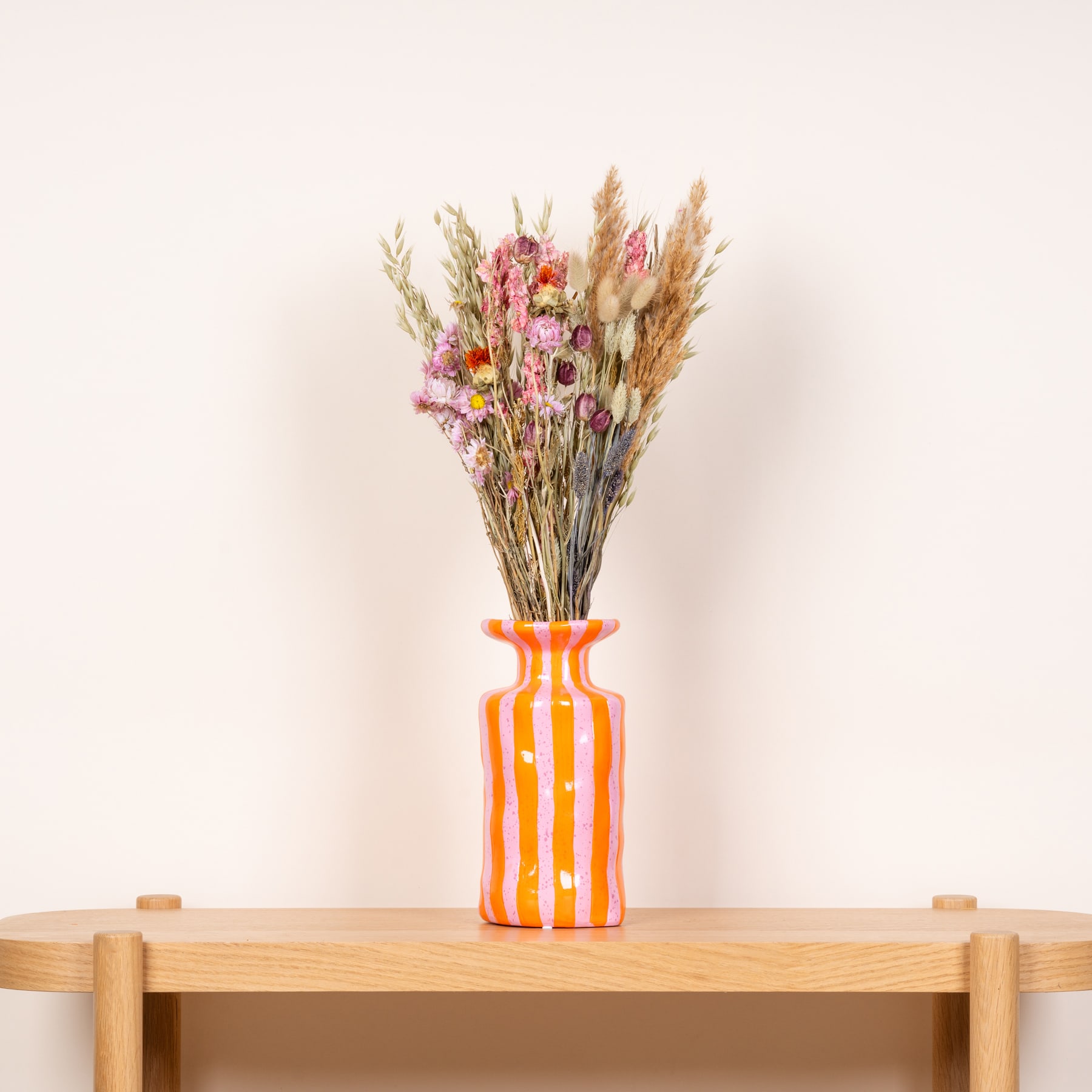 Ein farbenfroher Strauß getrockneter Blumen in einer orange-weiß gestreiften Vase steht auf einem Holztisch vor einem schlichten Hintergrund.
