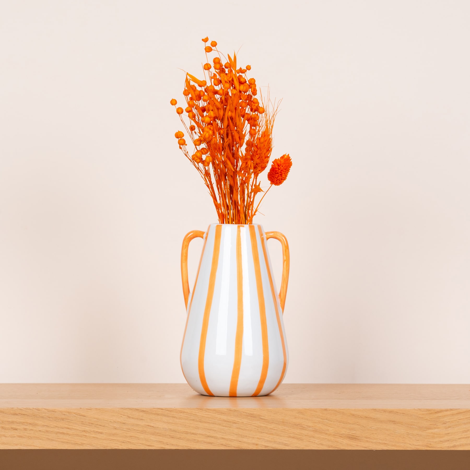 Eine weiß-orange Keramikvase gefüllt mit leuchtend orangefarbenen Trockenblumen steht auf einem Holztisch vor einem schlichten beigen Hintergrund.