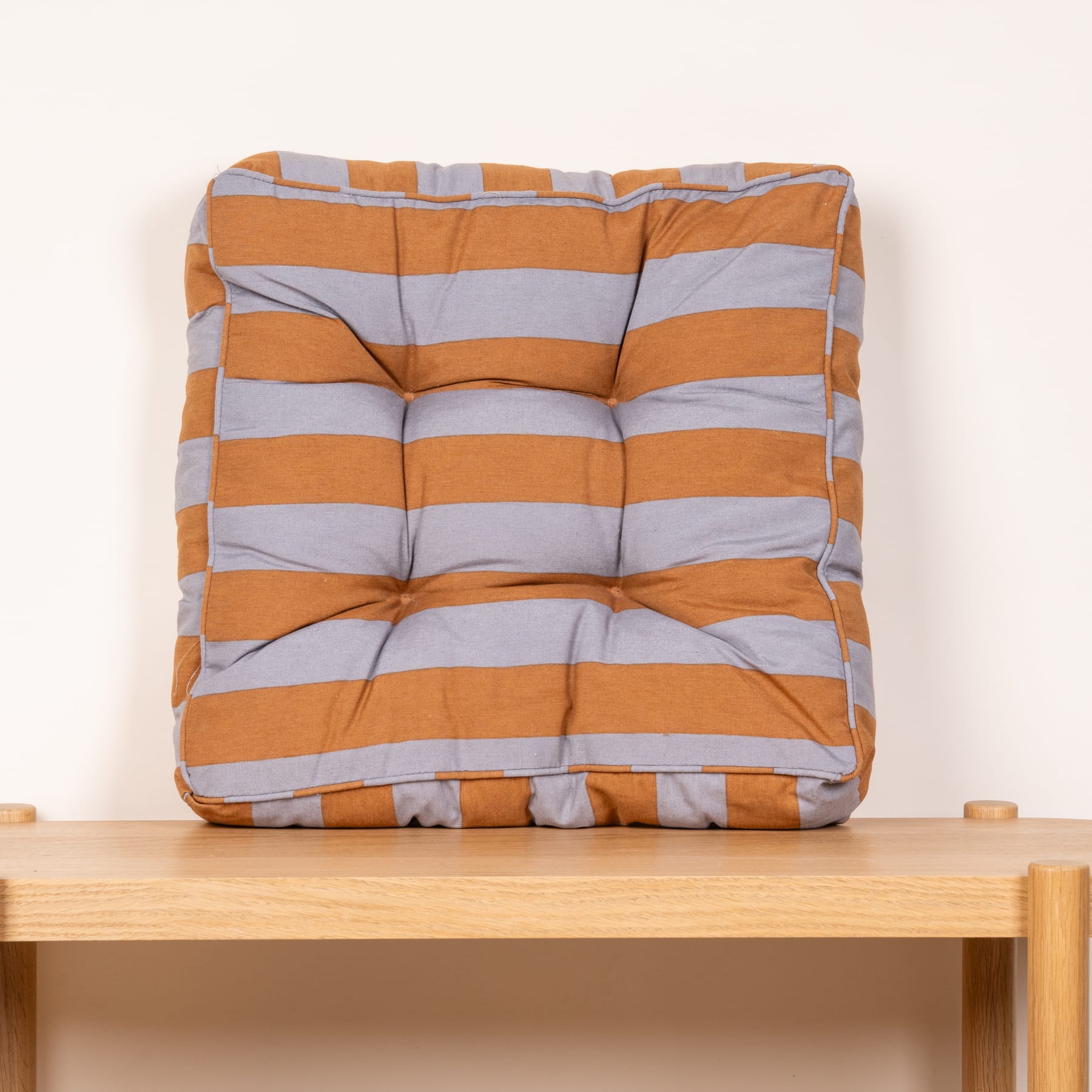 Ein gestreiftes orange-graues Kissen liegt auf einem Holzregal vor einem schlichten weißen Hintergrund.