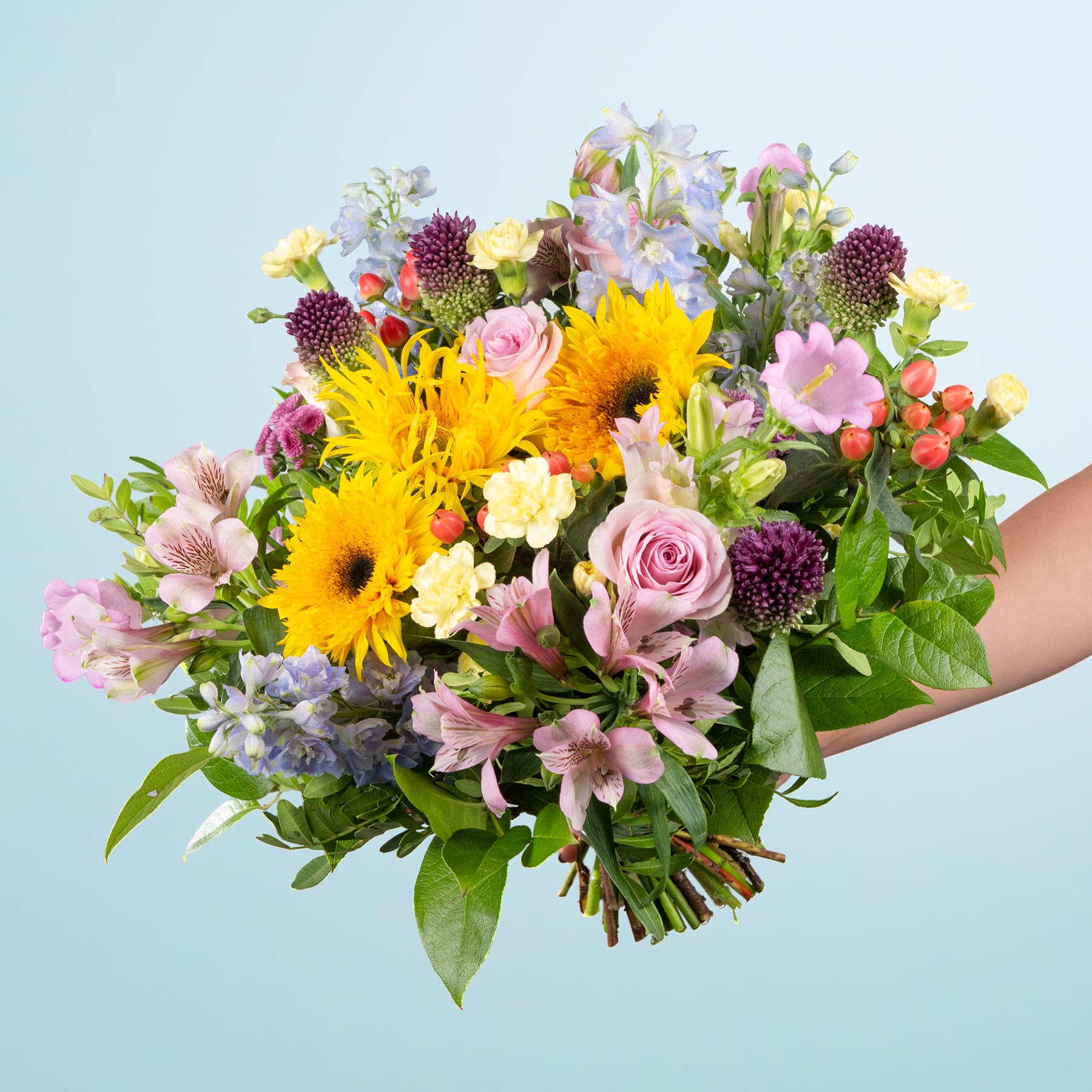 Ein leuchtender Strauß bunter Blumen, darunter Sonnenblumen, Rosen und Chrysanthemen, wird vor einem hellblauen Hintergrund in den Händen einer Person gehalten.