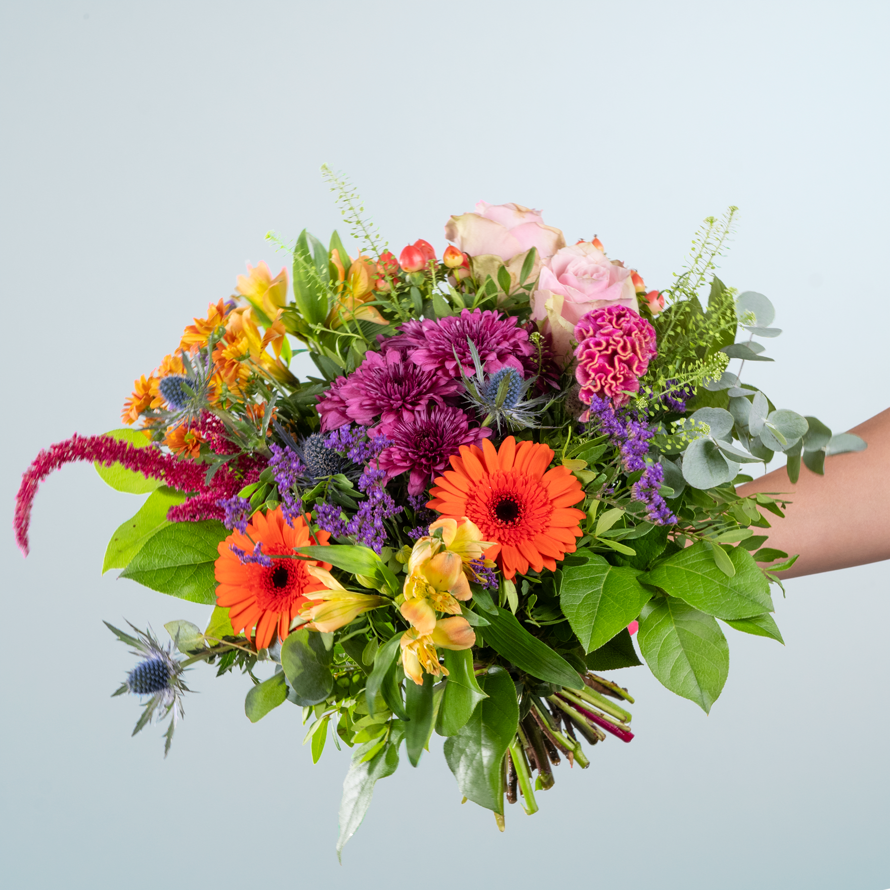 Ein leuchtender Strauß bunter Blumen, darunter Rosen, Gerbera und Chrysanthemen, den eine Person vor einem schlichten, hellen Hintergrund in den Händen hält.