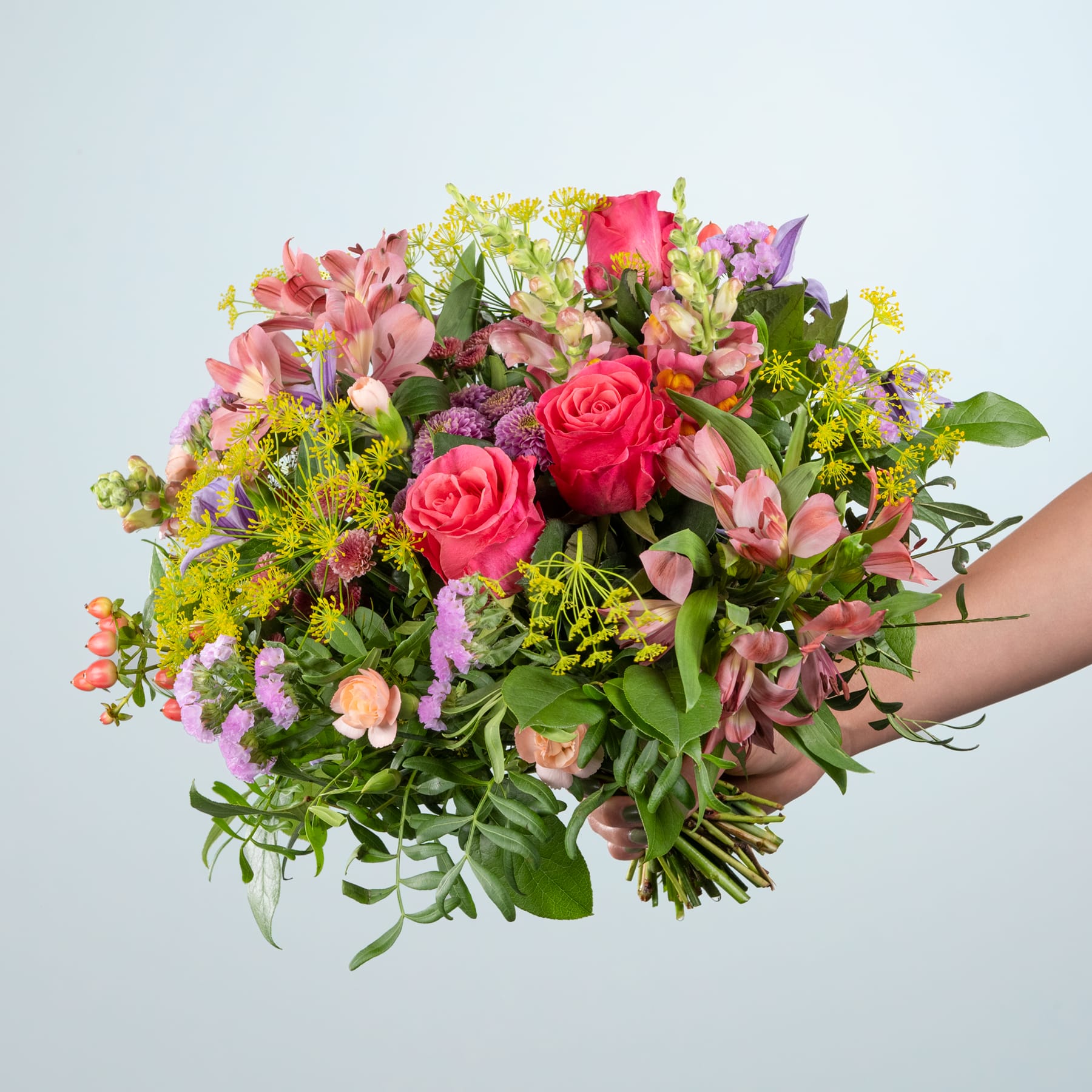 Ein leuchtender Strauß verschiedener bunter Blumen, darunter rote Rosen, rosa Inkalilien und gelbe Chrysanthemen, die eine Person vor einem schlichten hellen Hintergrund in den Händen hält.