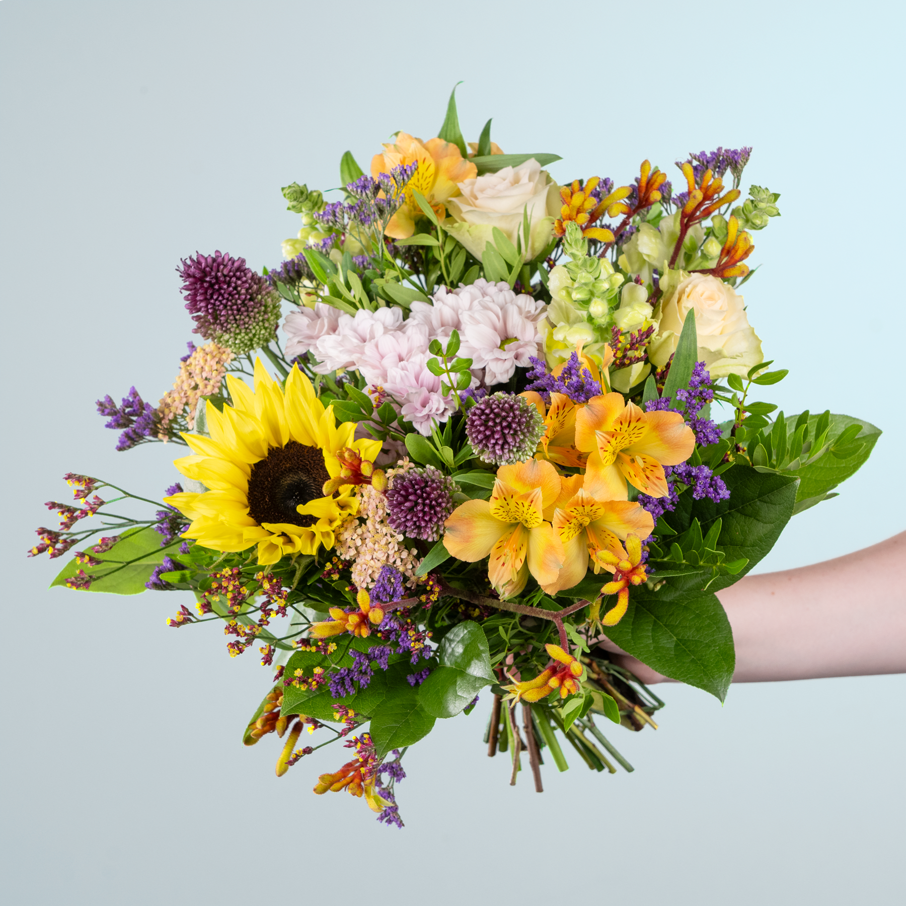 Ein leuchtender Strauß bunter Blumen, darunter Sonnenblumen, Dahlien und verschiedene andere Blüten, den eine Person vor einem schlichten Hintergrund in den Händen hält.