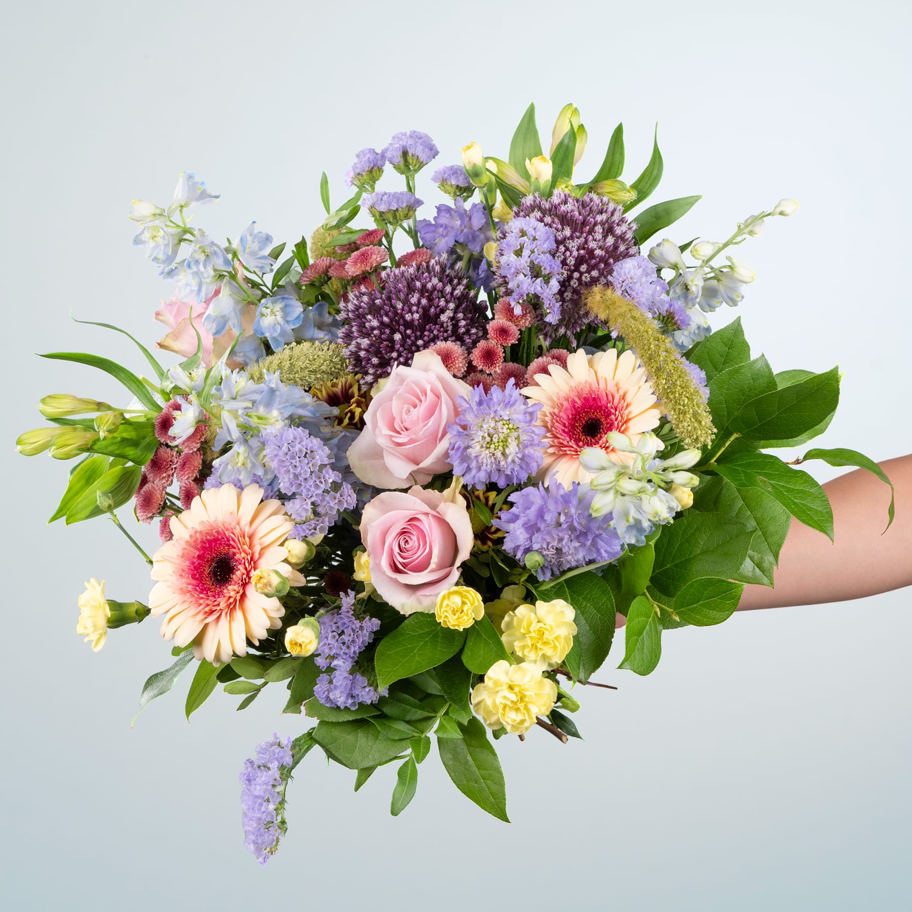 Ein lebendiger Strauß verschiedener bunter Blumen, darunter rosa Rosen, violette Hyazinthen, gelbe Narzissen und rosa und weiße Gerbera, vor einem schlichten hellgrauen Hintergrund.