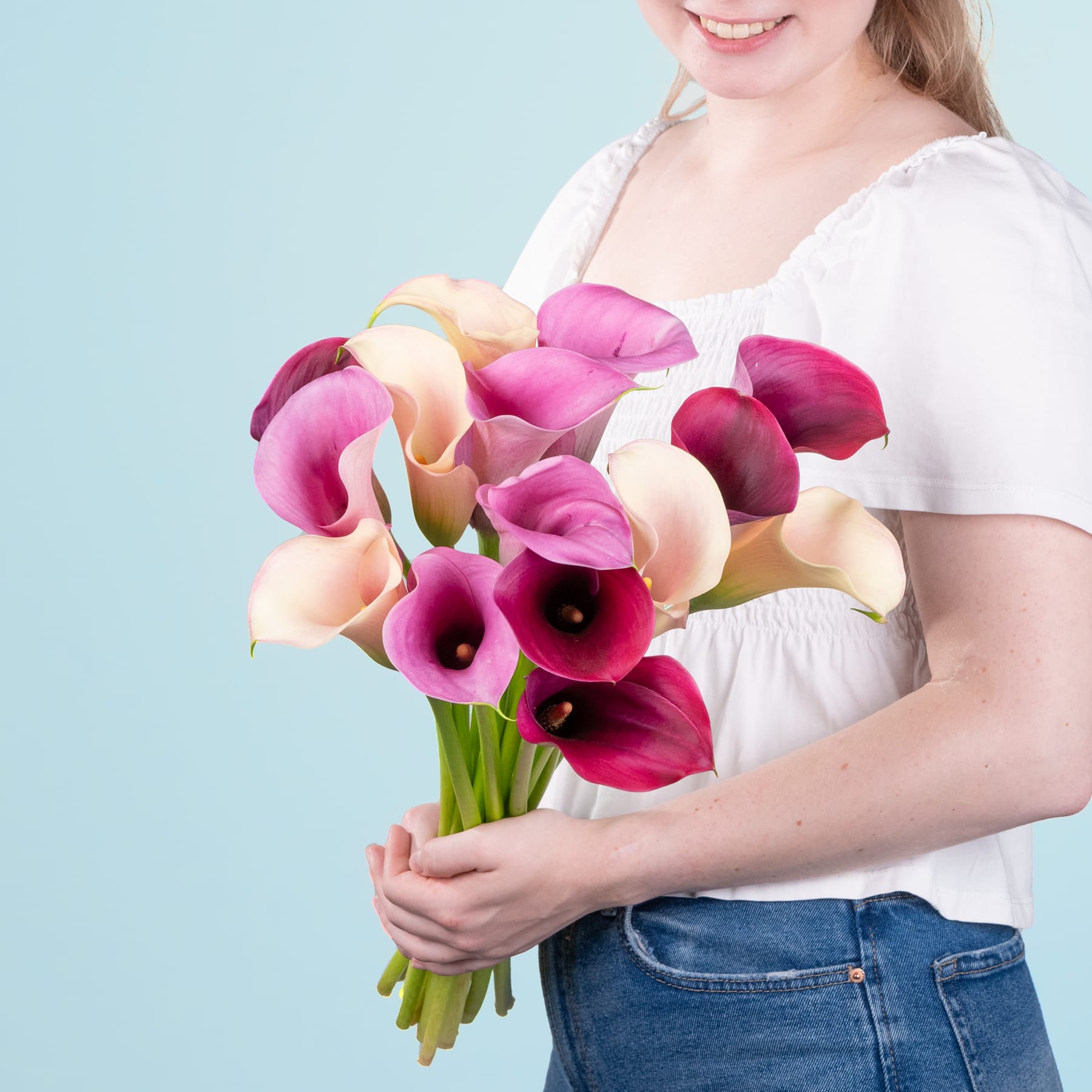 Eine lächelnde junge Frau hält einen farbenfrohen Strauß rosa und gelber Calla-Lilien vor einem hellblauen Hintergrund.