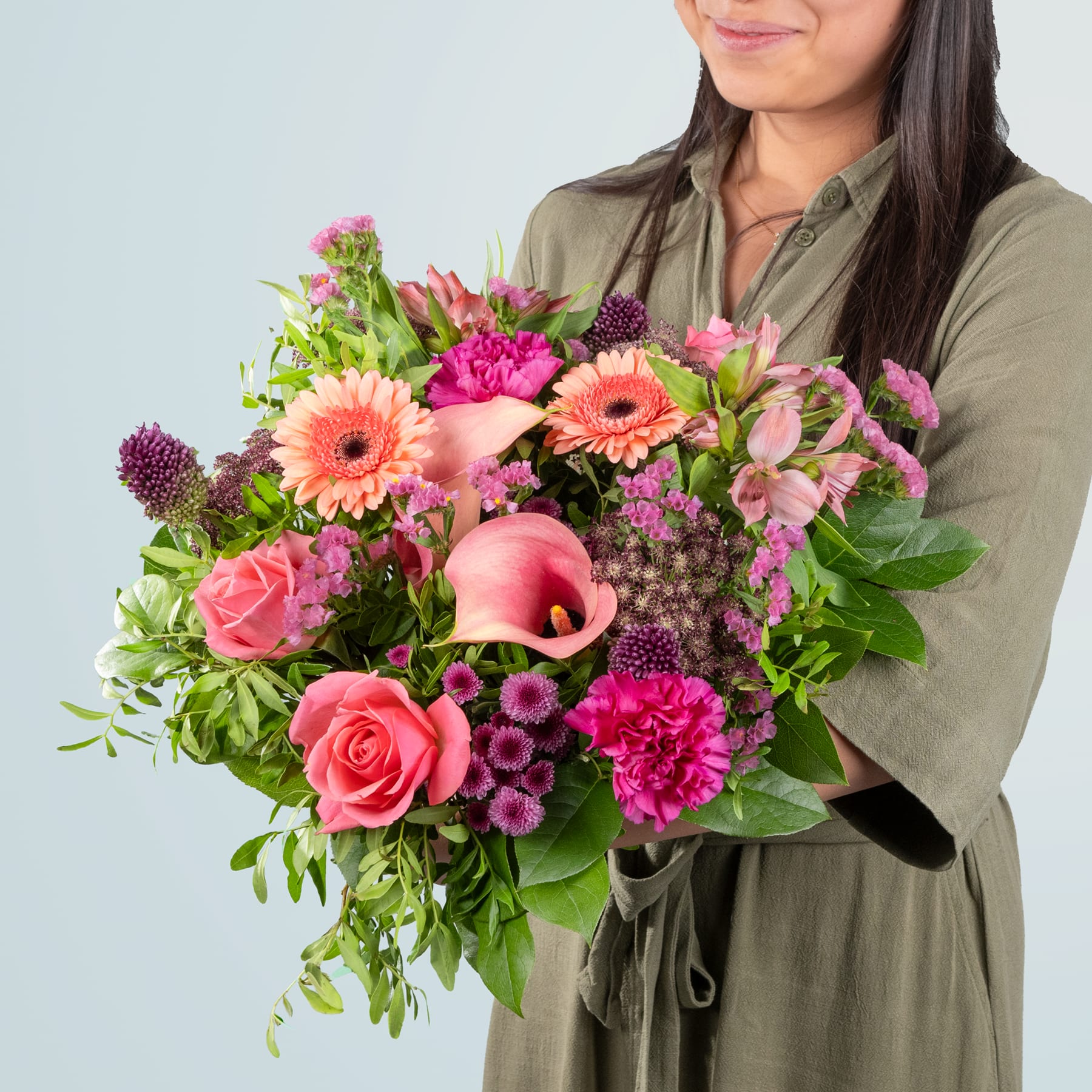 Ein leuchtender Strauß bunter Blumen, darunter Rosen, Gerbera und Nelken, wird von einer Person in einer grünen Jacke vor einem schlichten Hintergrund gehalten.