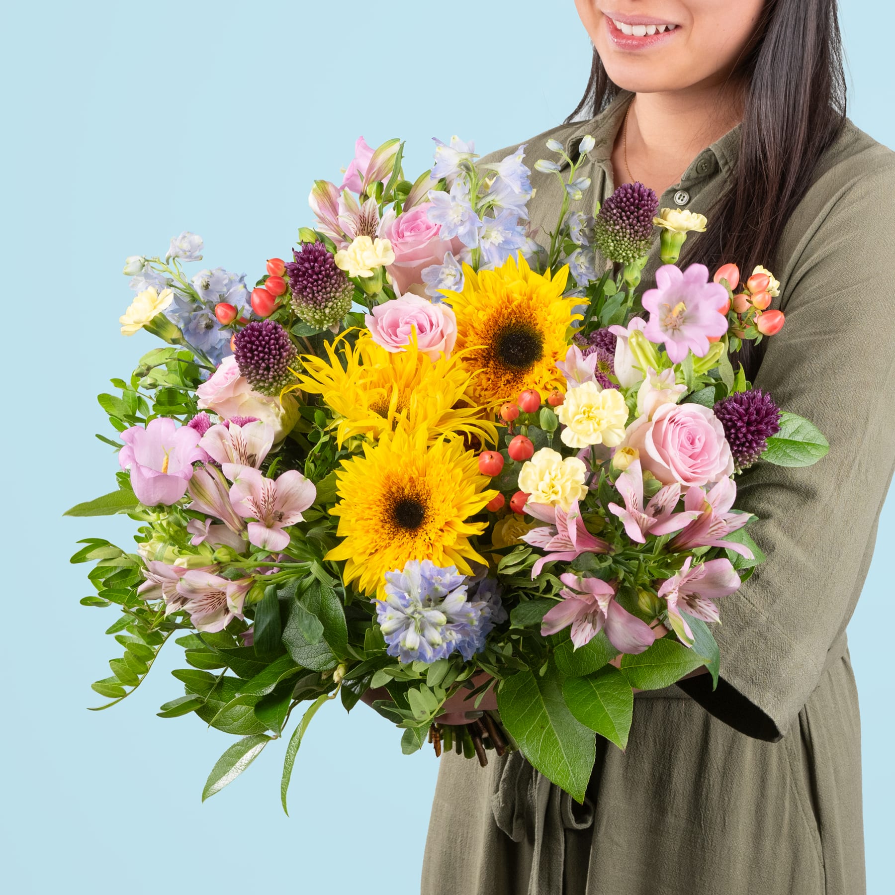Ein leuchtender Strauß bunter Blumen, darunter Sonnenblumen, Rosen und andere Blüten, wird von einer lächelnden Person in einer grünen Jacke vor einem hellblauen Hintergrund gehalten.