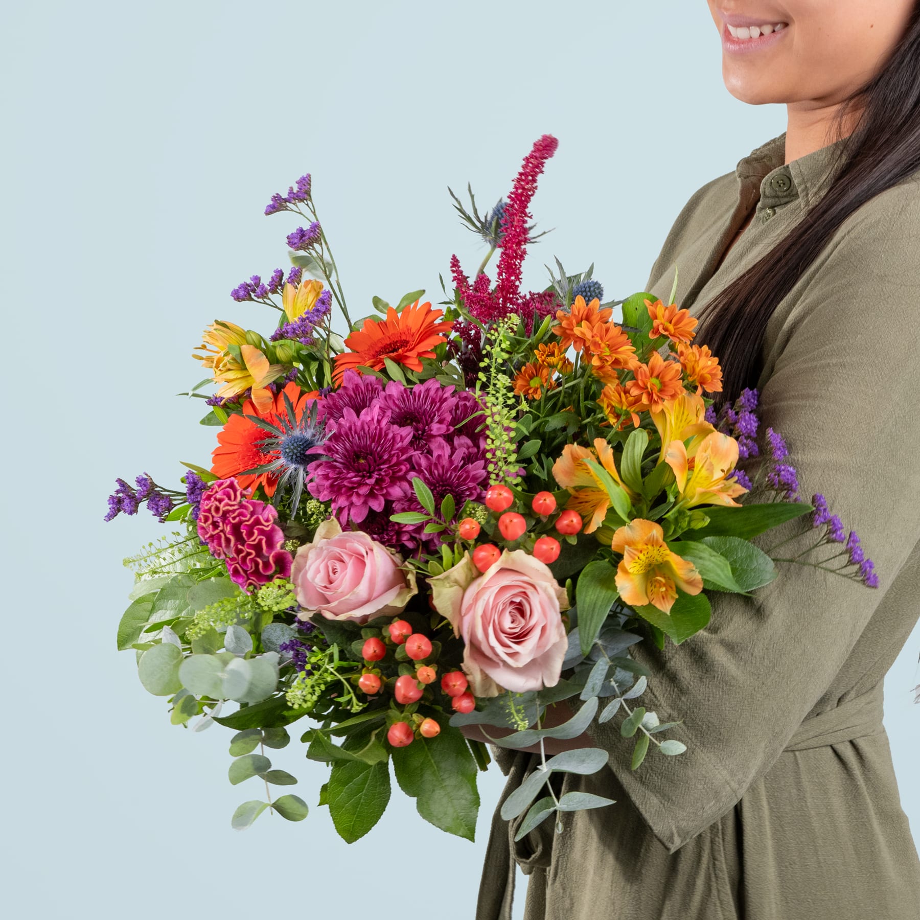 Ein leuchtender Strauß bunter Blumen, darunter Rosen, Gänseblümchen und Chrysanthemen, wird von einer Person in einem beigen Mantel vor einem schlichten hellblauen Hintergrund gehalten.