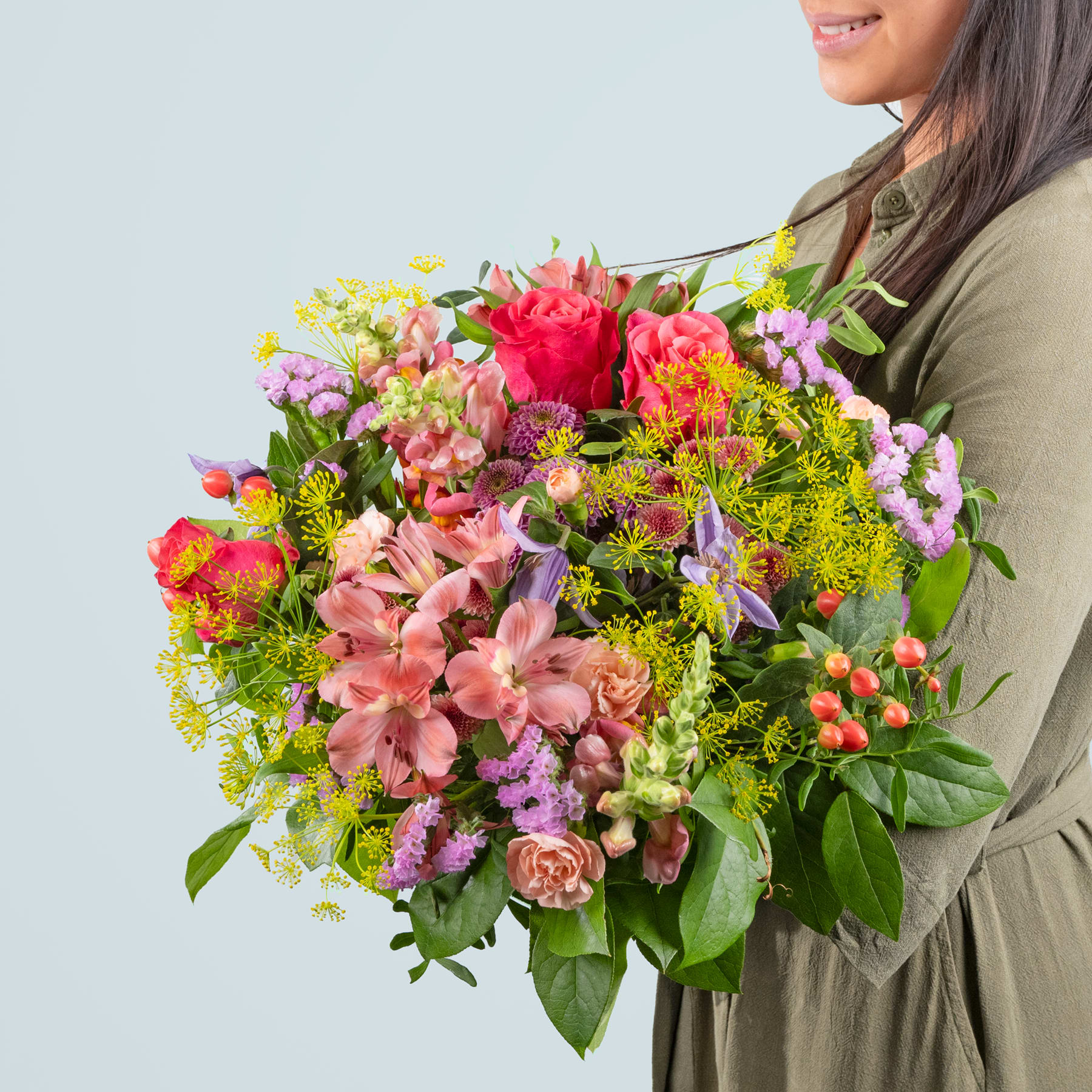 Ein leuchtender Strauß bunter Blumen, darunter Rosen, Lilien und andere Blüten, wird von einer Person in einer grünen Jacke vor einem schlichten Hintergrund gehalten.