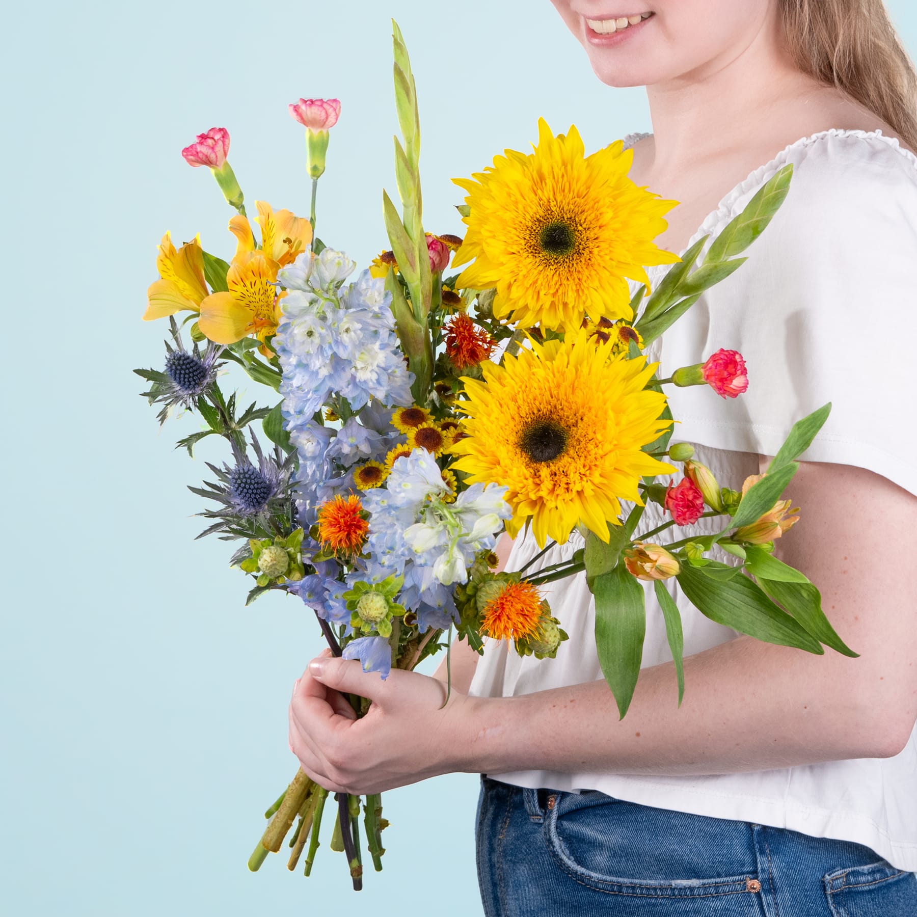 Eine junge Frau in einem weißen Hemd hält einen farbenfrohen Strauß aus Sonnenblumen, Gänseblümchen und anderen bunten Blumen vor einem hellblauen Hintergrund.