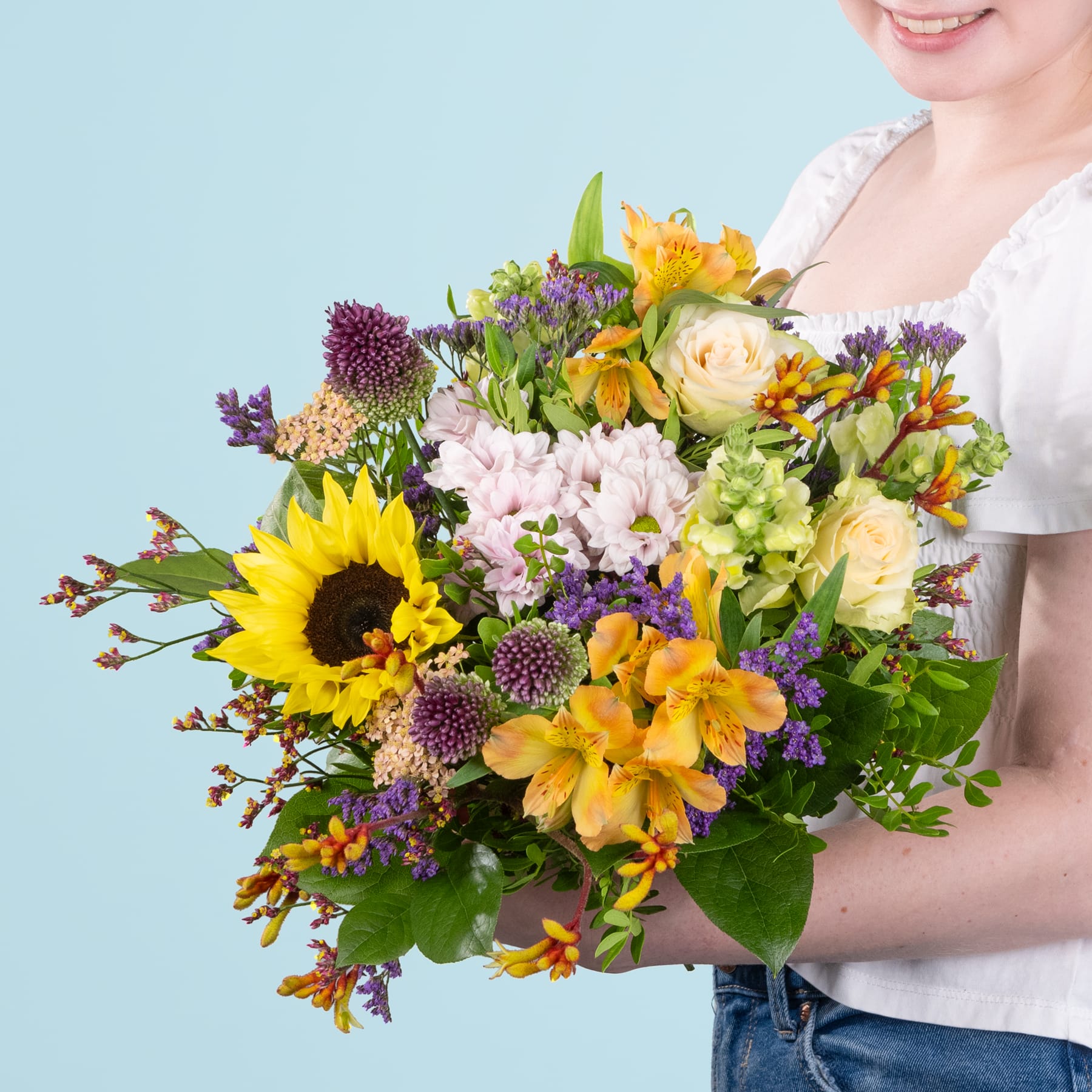 Ein leuchtender Strauß bunter Blumen, darunter Sonnenblumen, Rosen und verschiedene andere Blüten, wird von einer Person im Vordergrund vor einem hellblauen Hintergrund gehalten.