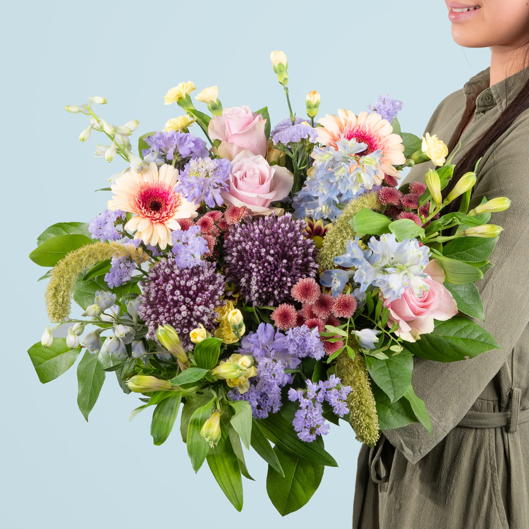 Ein leuchtender Strauß bunter Blumen, darunter Rosen, Gänseblümchen und Flieder, wird von einer Person gehalten, die einen Trenchcoat vor einem schlichten blauen Hintergrund trägt.