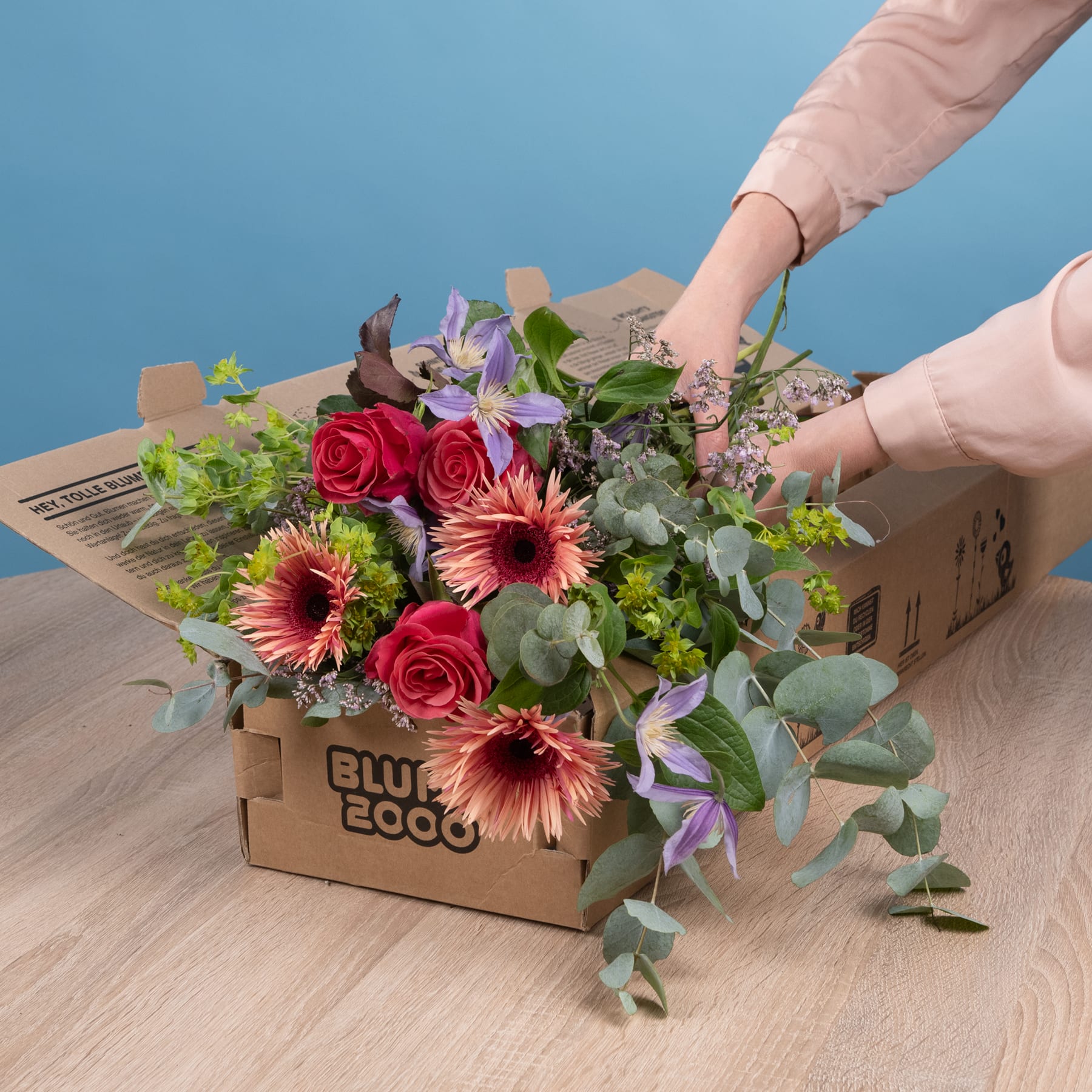 Ein farbenfroher Strauß roter Rosen, rosa Gerbera und violetter Blumen steht in einem Karton auf einer Holzoberfläche vor blauem Hintergrund.