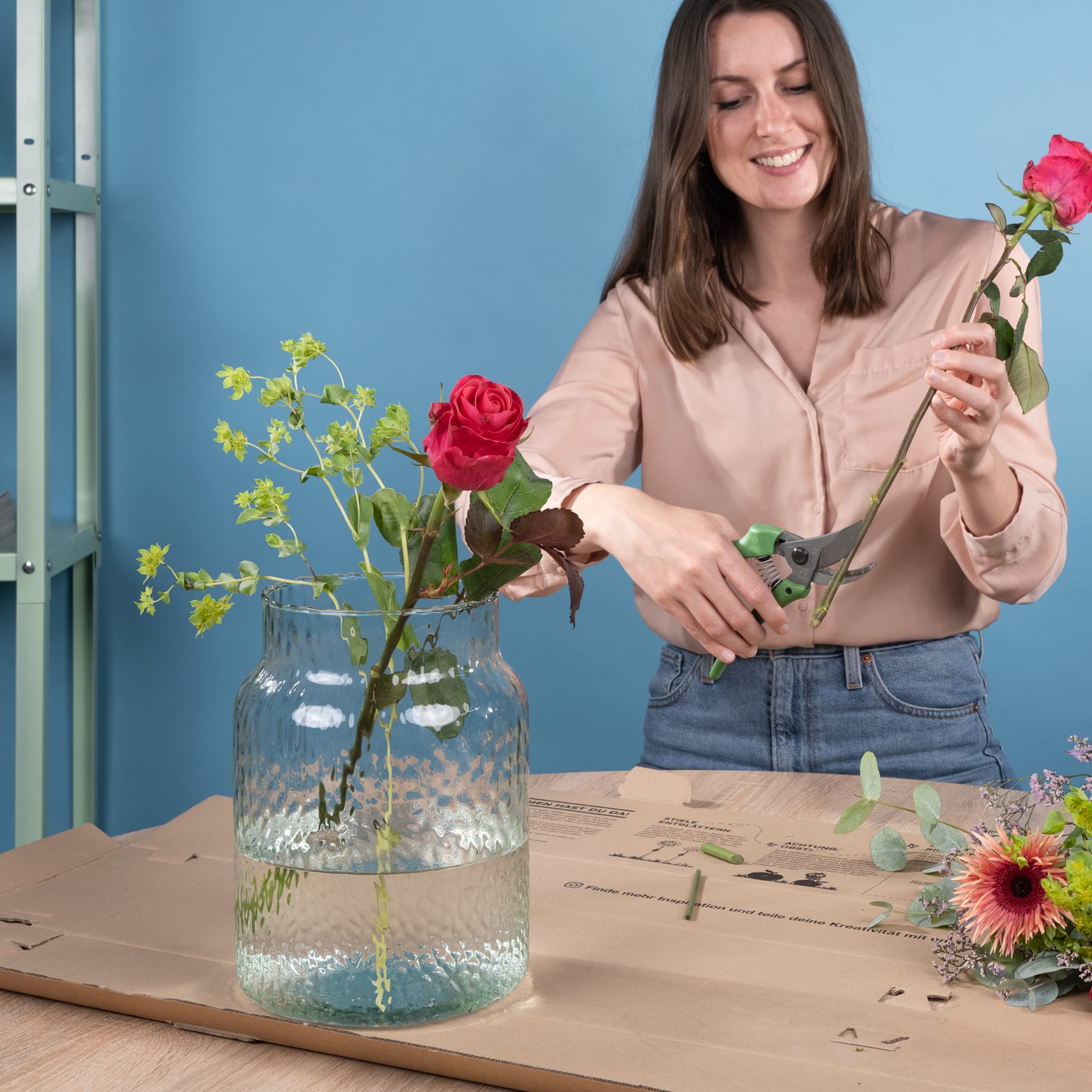 Eine junge Frau mit langen braunen Haaren arrangiert einen Strauß bunter Blumen, darunter eine rote Rose, in einer Glasvase auf einem Holztisch vor blauem Hintergrund.