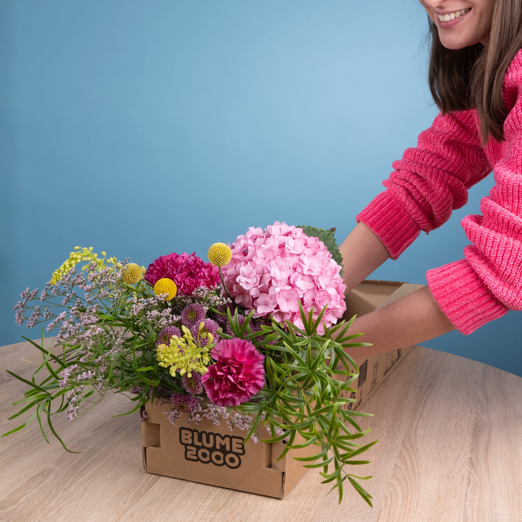 Ein farbenfroher Strauß aus rosa Hortensien, roten Dahlien und gelben Chrysanthemen wird in einem Karton auf einer Holzoberfläche vor blauem Hintergrund arrangiert.