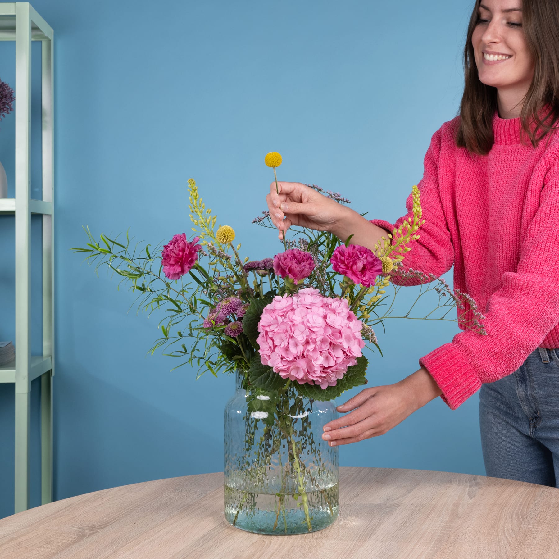Eine Frau in einem rosa Pullover arrangiert einen Strauß leuchtend rosa und gelber Blumen in einer Glasvase auf einem Holztisch vor einem blauen Hintergrund.