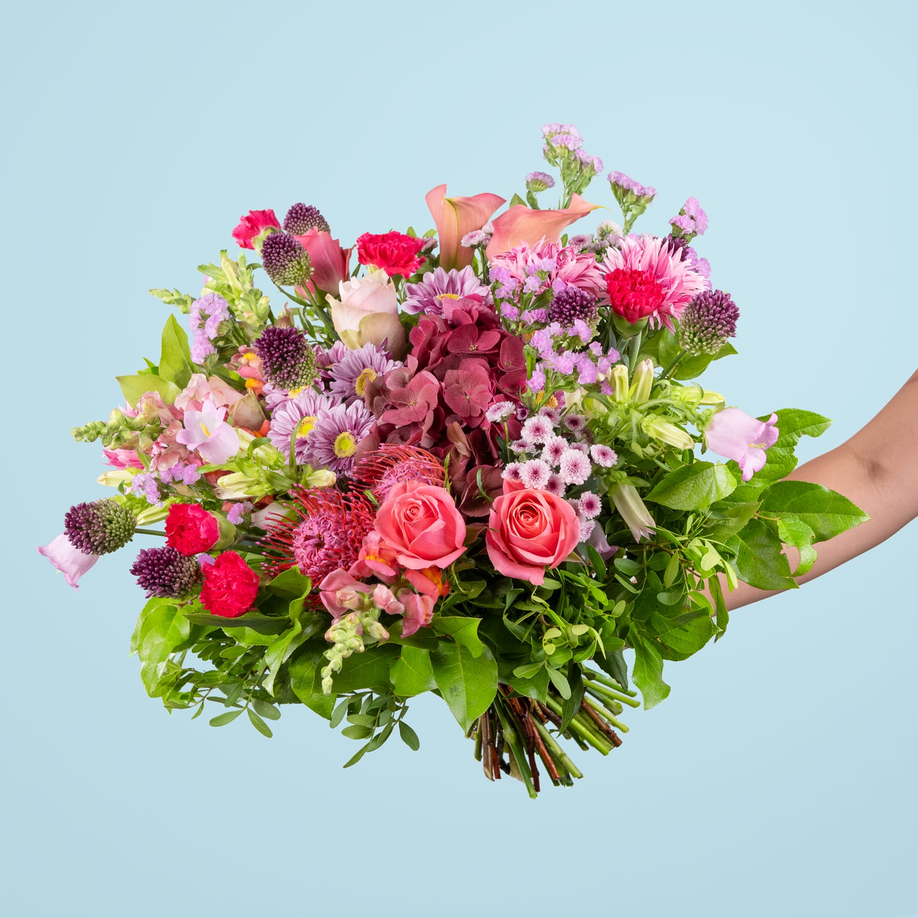 Ein lebendiger und üppiger Strauß verschiedener bunter Blumen, darunter Rosen, Nelken und Chrysanthemen, den eine Person vor einem schlichten blauen Hintergrund in den Händen hält.