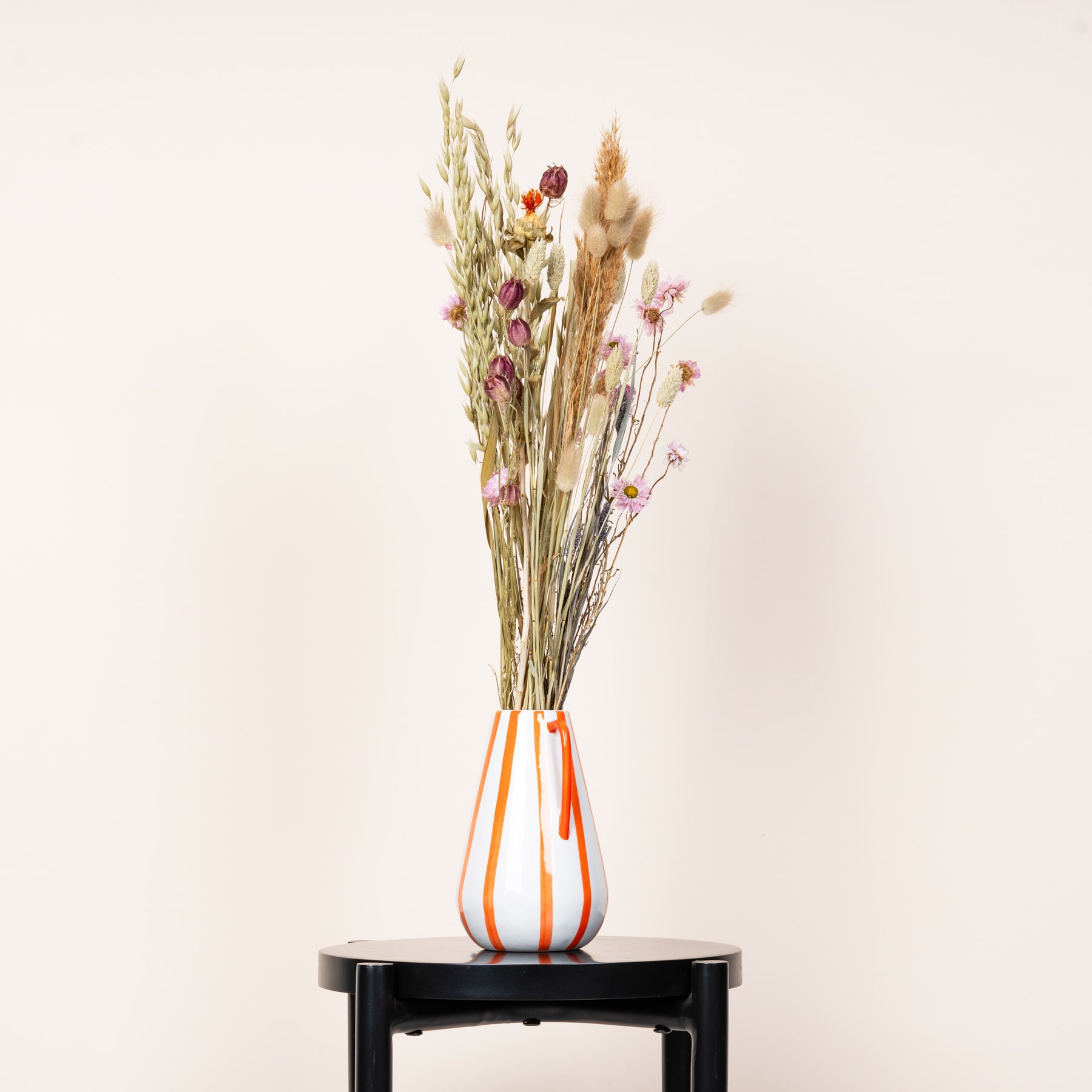 Ein Strauß getrockneter Blumen, darunter Weizenhalme und rosa Blüten, wird in einer Vase mit bunten Streifen vor einem schlichten, hellen Hintergrund präsentiert.