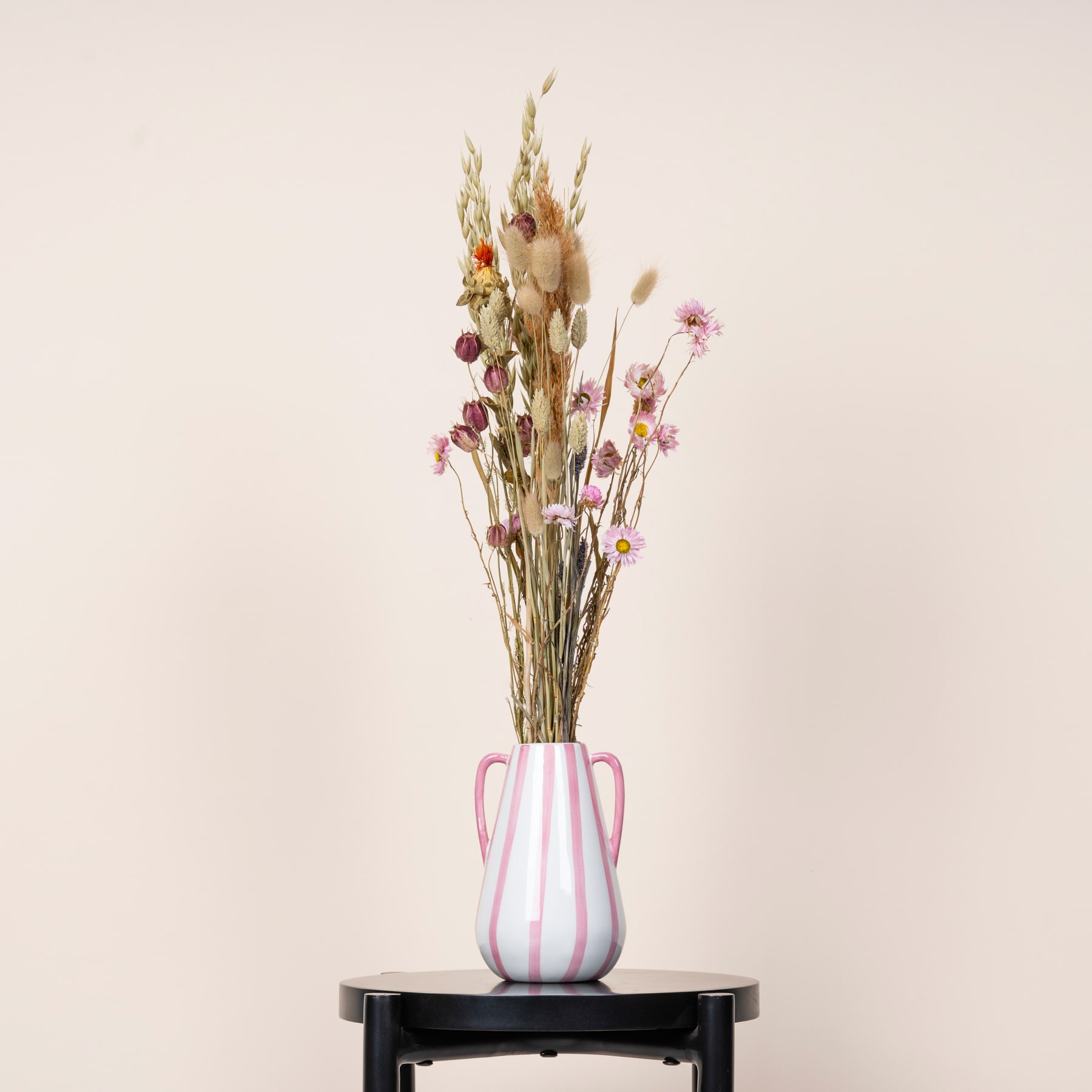 Eine Vase mit einem Strauß getrockneter Blumen, darunter Weizenhalme und rosa Blüten, steht auf einer dunklen Oberfläche vor einem schlichten, hellen Hintergrund.