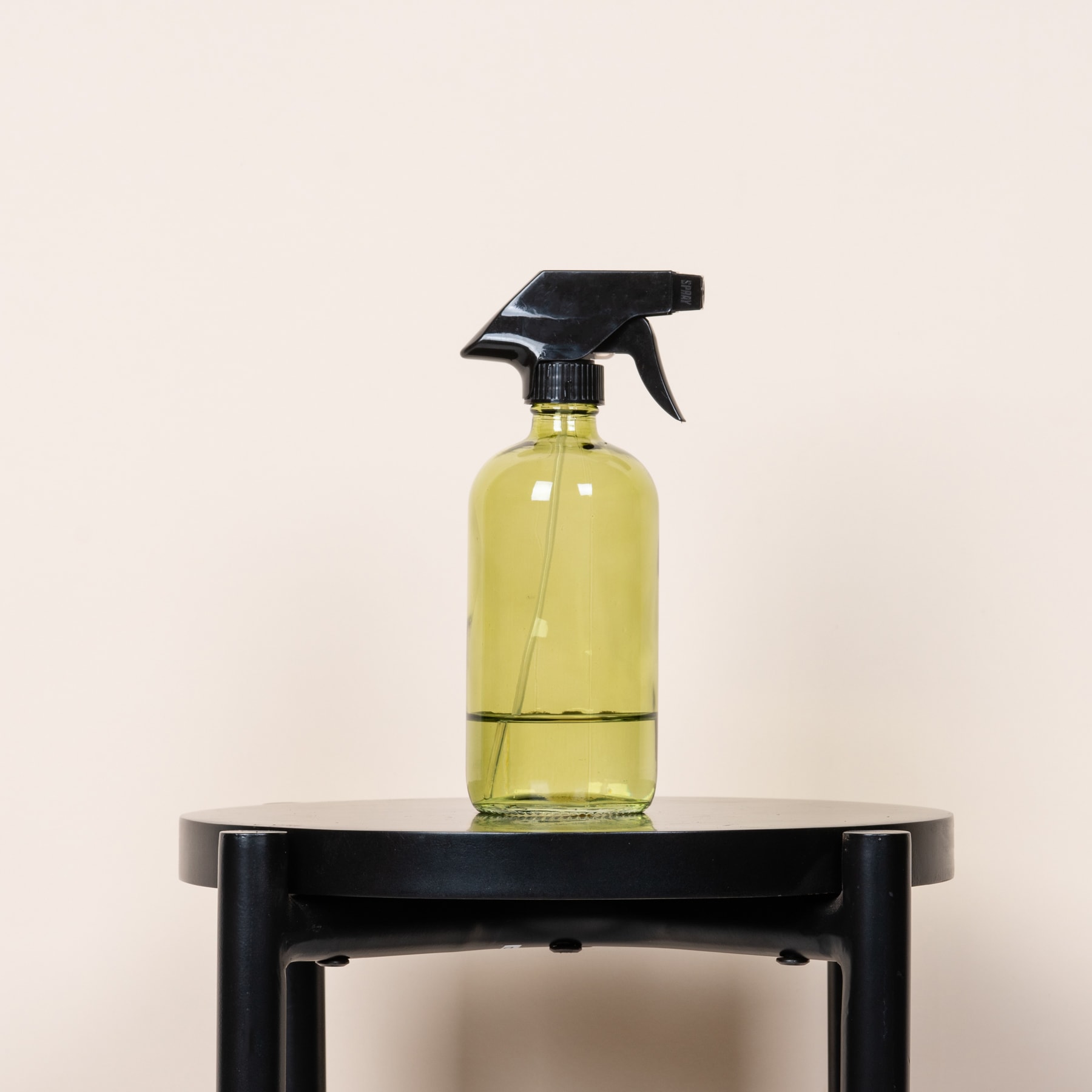 Eine gelbe Sprühflasche mit schwarzem Auslöser steht auf einer schwarzen Plattform vor einem schlichten beigen Hintergrund.