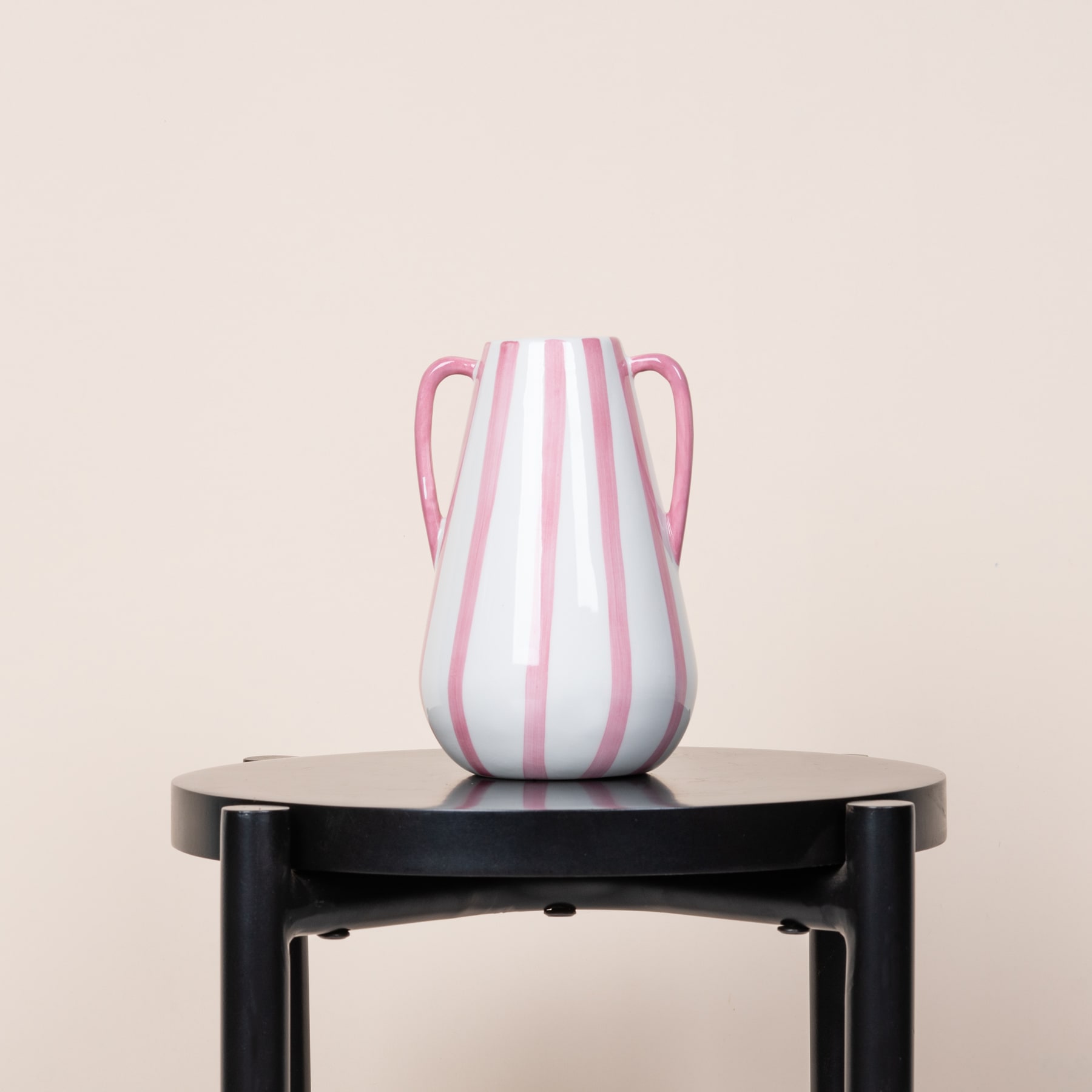 Eine rosa-weiß gestreifte Keramikvase steht auf einer schwarzen runden Plattform vor einem hellbeigen Hintergrund.