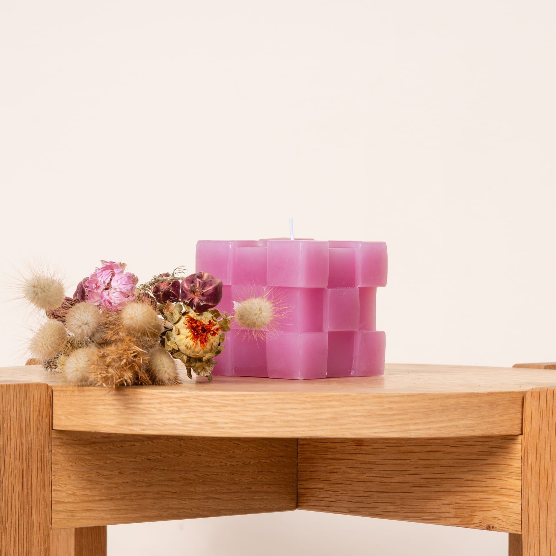 Das Bild zeigt ein Holzregal mit einem Strauß rosa und lila Blumen und einem Satz rosa würfelförmiger Kerzen.