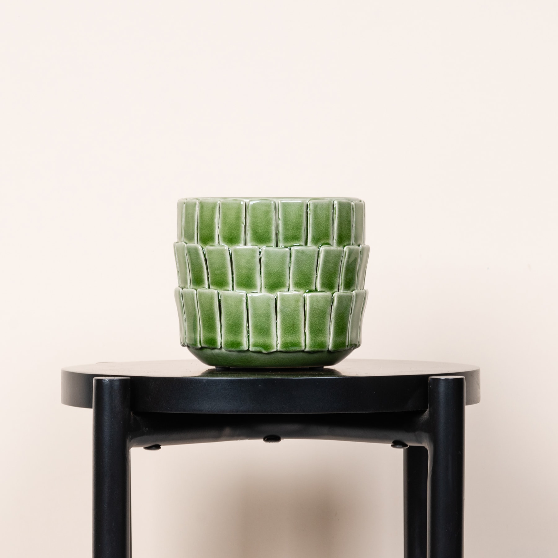 Ein grüner Blumentopf oder eine Vase aus Keramik steht auf einem schwarzen Metallständer vor einem schlichten, hellen Hintergrund.