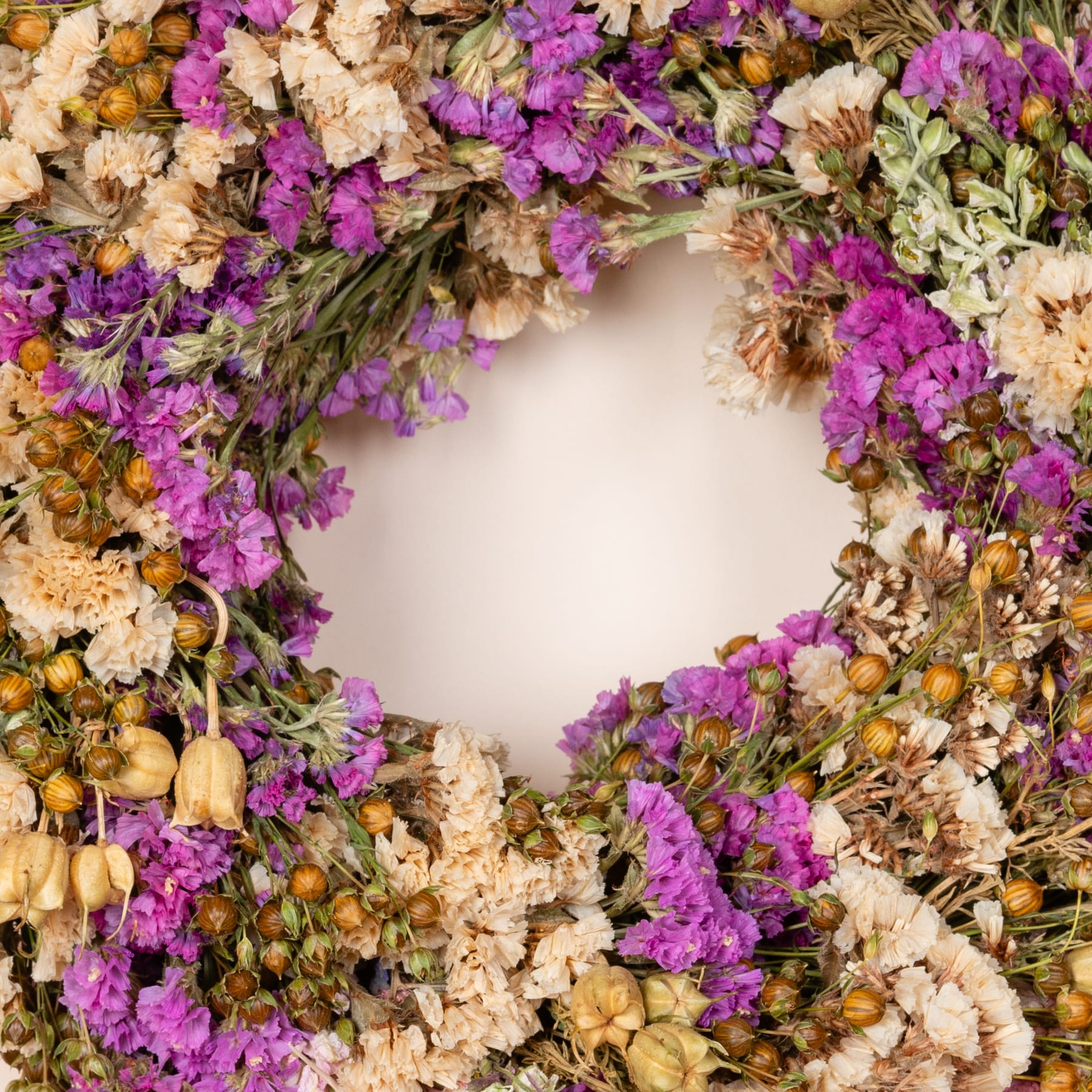 Ein kranzartiges Arrangement aus leuchtend violetten Blüten und getrockneten Blättern und Stängeln, das vor einem neutralen Hintergrund eine warme und natürliche Komposition schafft.