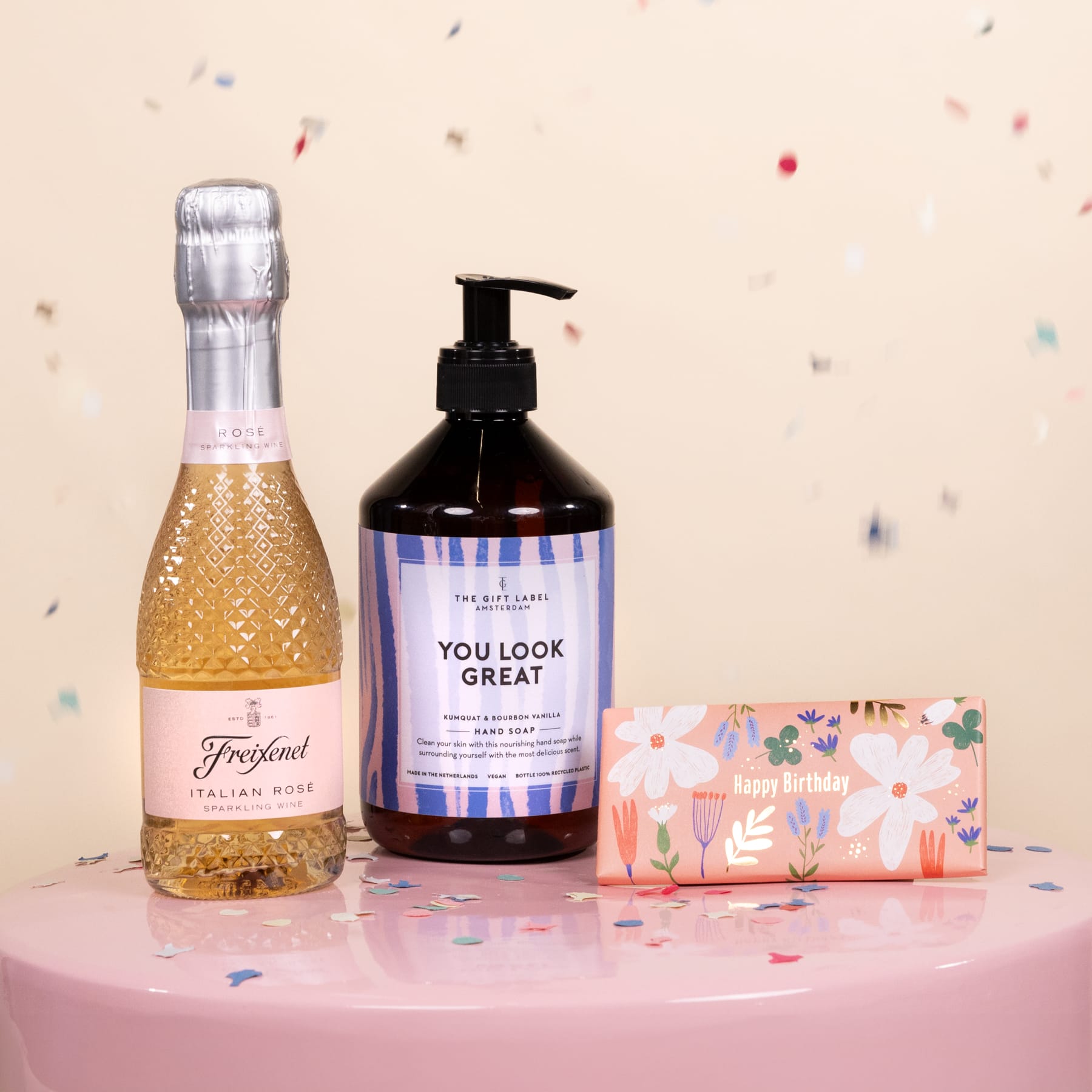 Ein rosa Tisch mit einer Flasche Sekt, einer Flasche Handseife und einer Geschenkbox mit Blumenmuster, alles vor einem hellen Hintergrund mit verstreutem Konfetti.