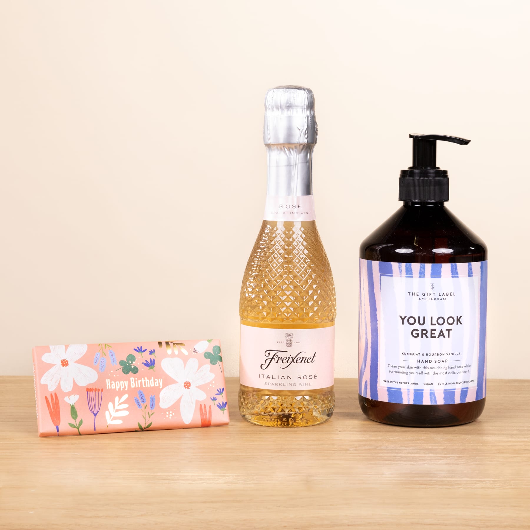 Eine Flasche Sekt, eine Flasche Handseife und eine Geschenkbox mit floralen Mustern sind auf einer Holzoberfläche vor einem hellen Hintergrund arrangiert.