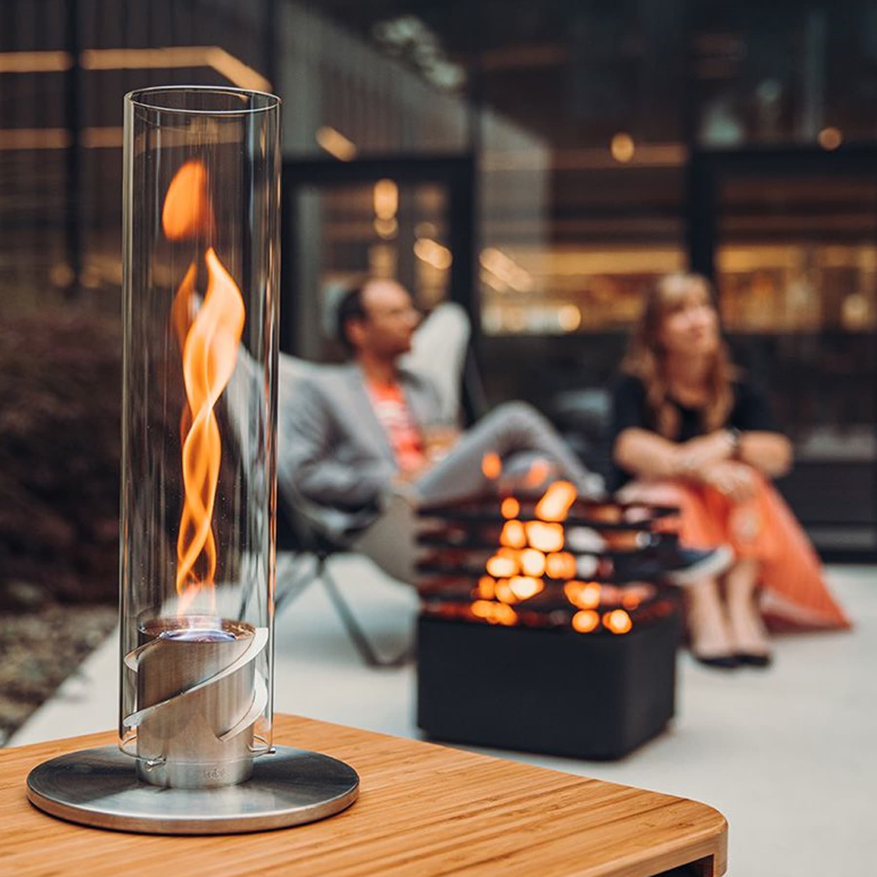 Ein Glaszylinder mit flackernder Flamme steht auf einer Holzoberfläche, während sich im Hintergrund Menschen um einen Kamin versammelt haben und eine gemütliche und einladende Atmosphäre schaffen.
