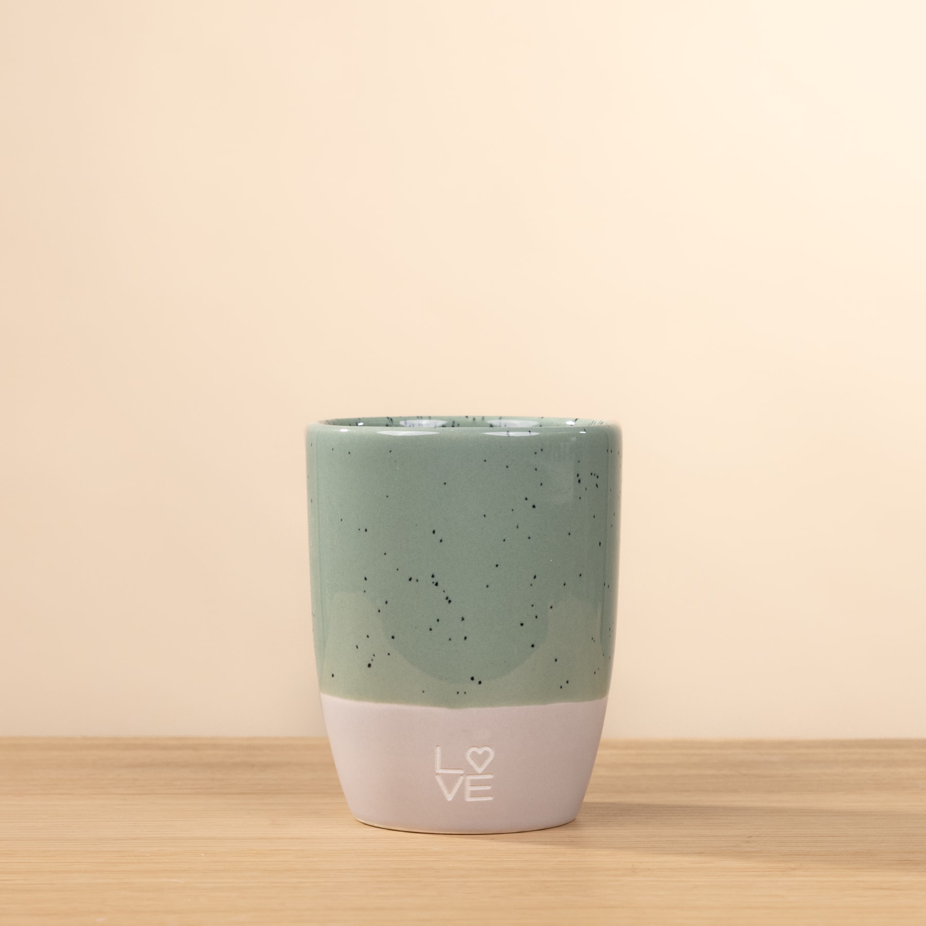 Eine Keramiktasse mit gesprenkelter mintgrüner Außenseite und weißem Boden, die auf einer Holzoberfläche vor einem hellbeigen Hintergrund ruht.