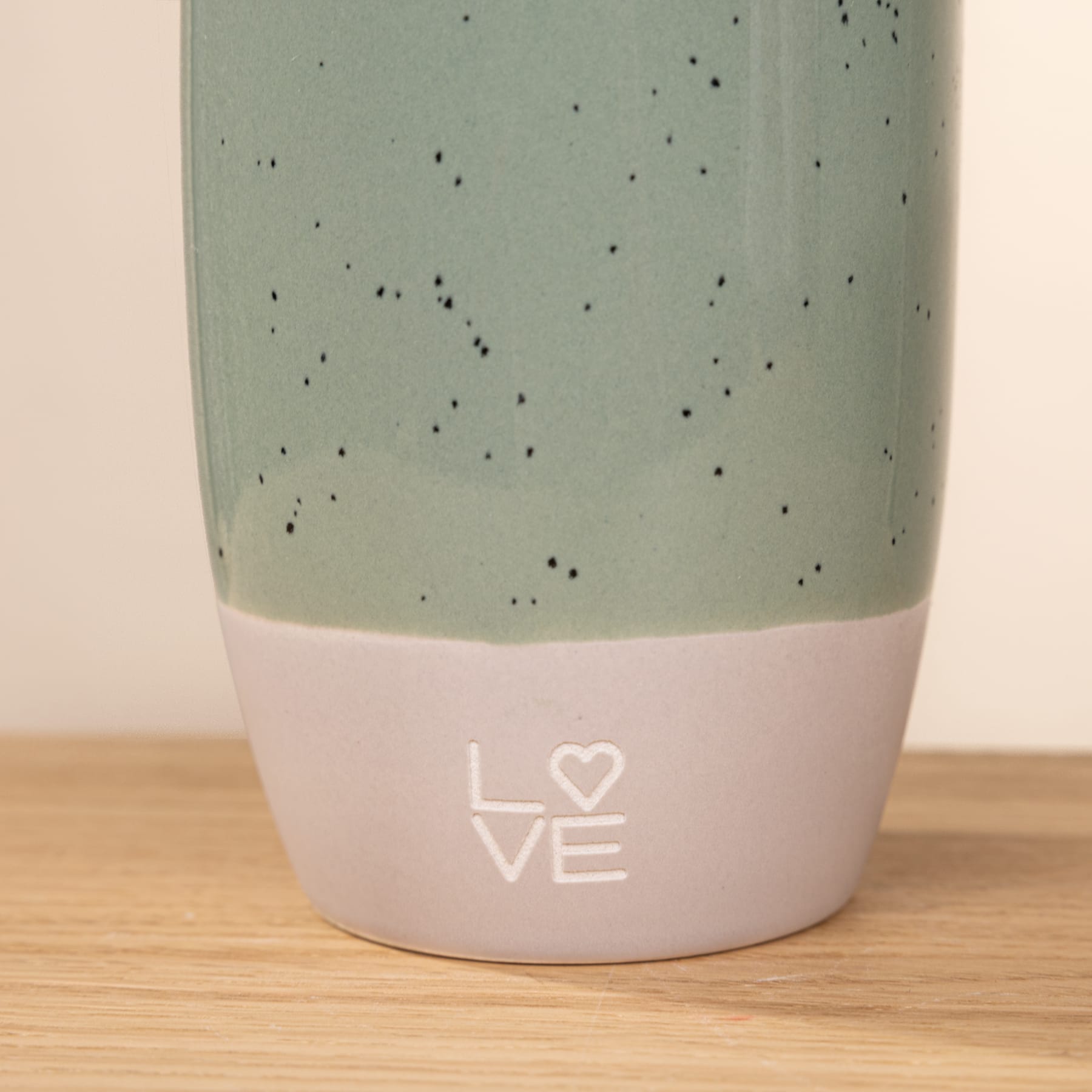 Ein Keramikbecher oder eine Tasse mit einer gesprenkelten mintgrünen Glasur und dem in Weiß geschriebenen Wort „LOVE“ im unteren Teil, ruht auf einer Holzoberfläche.