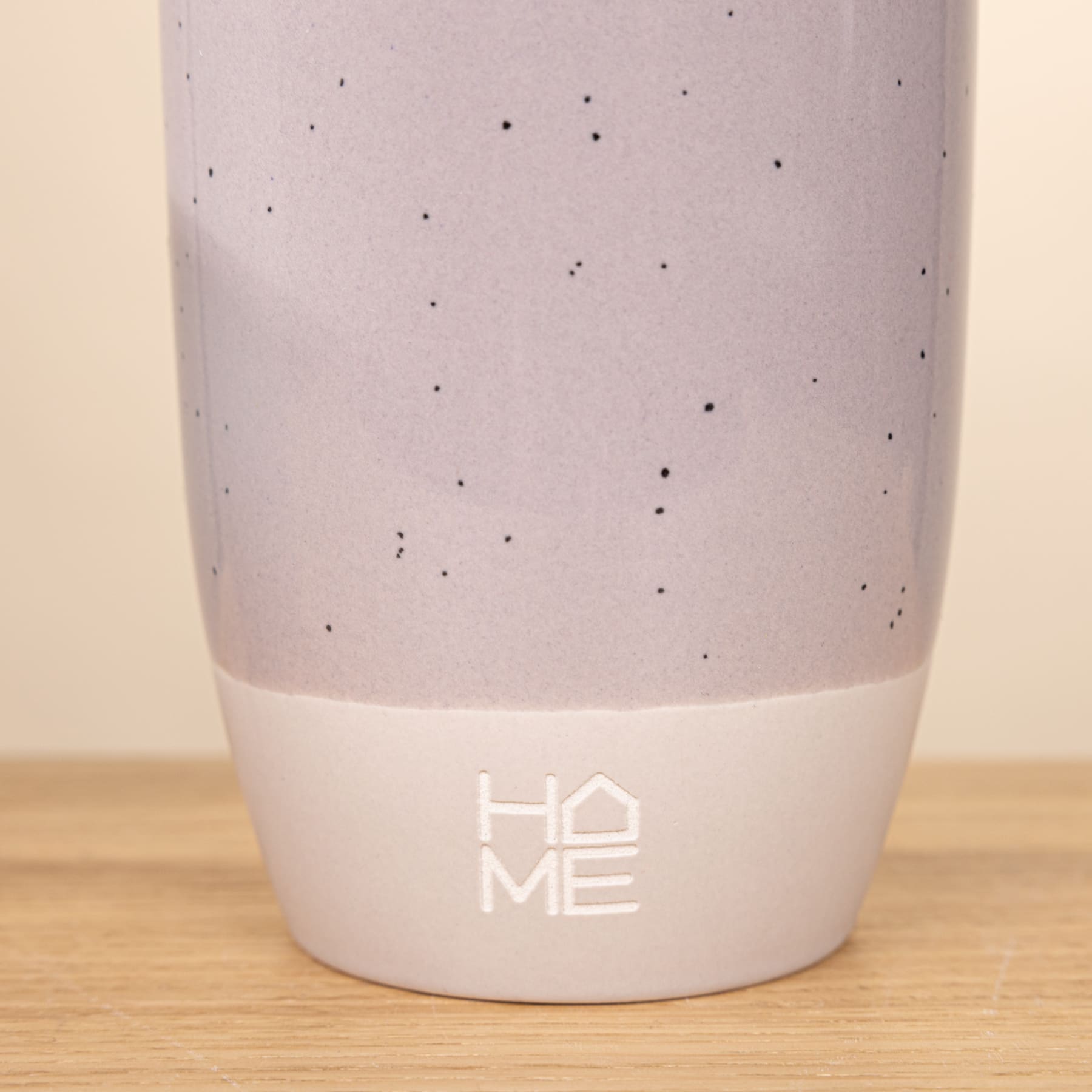 Eine weiße Keramiktasse oder ein Becher mit einem einfachen, minimalistischen Design mit kleinen schwarzen Punkten und dem aufgedruckten Text „HOME“ auf der Oberfläche, ruht auf einer Holzoberfläche.