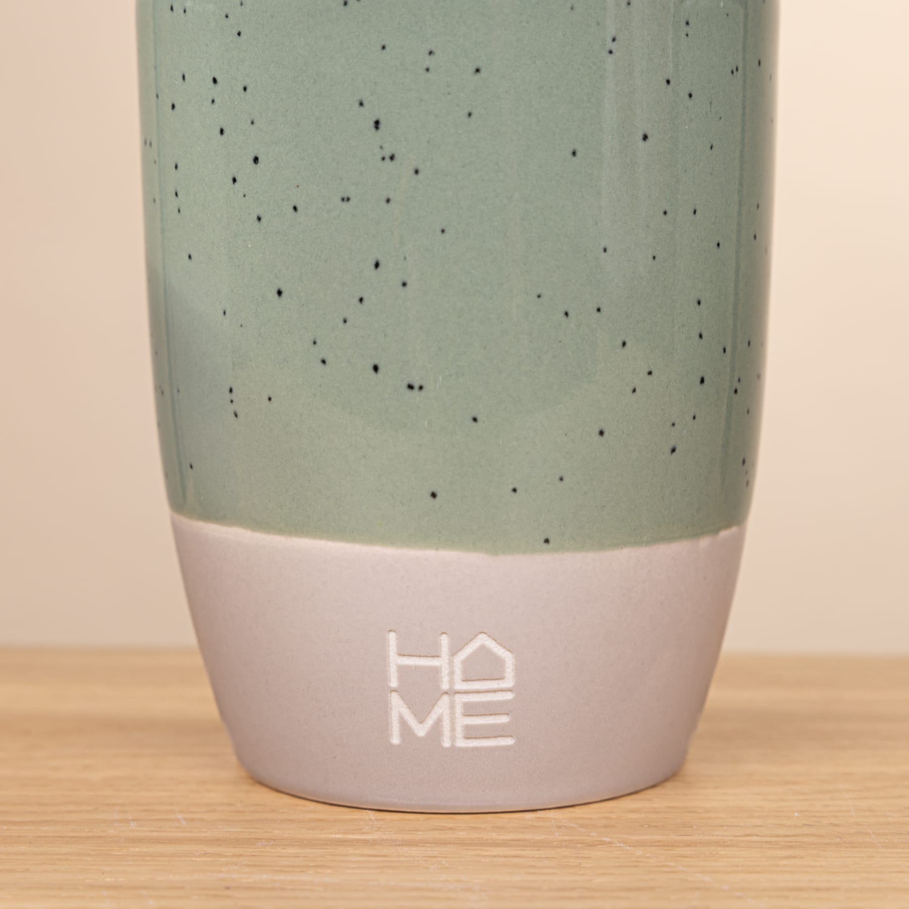 Eine Keramiktasse mit gesprenkelter mintgrüner Außenseite und einem weißen Band am Boden, auf der der Text „HOME“ aufgedruckt ist, steht auf einer Holzoberfläche.
