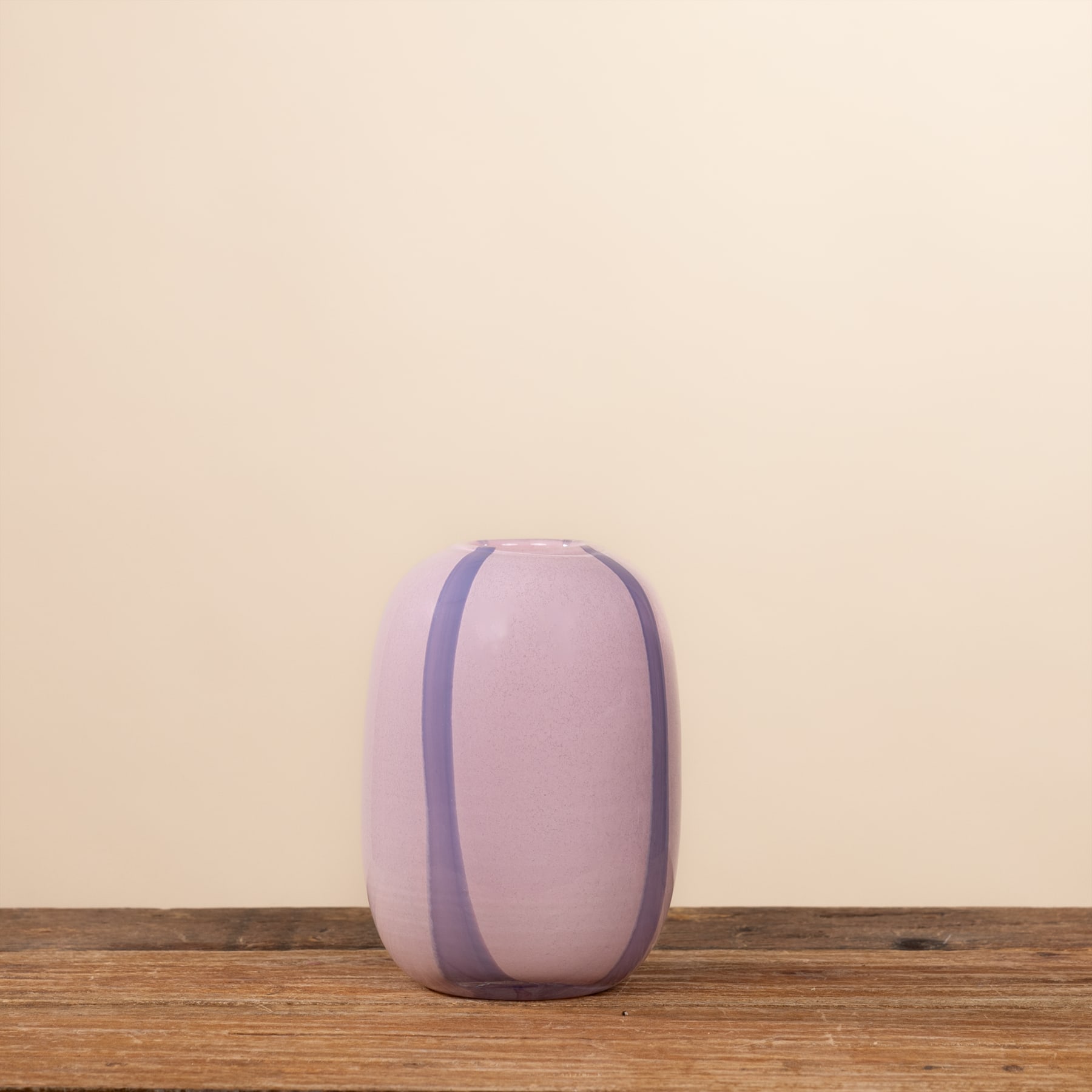 Eine lavendelfarbene Vase steht auf einer Holzoberfläche vor einem beigen Hintergrund.