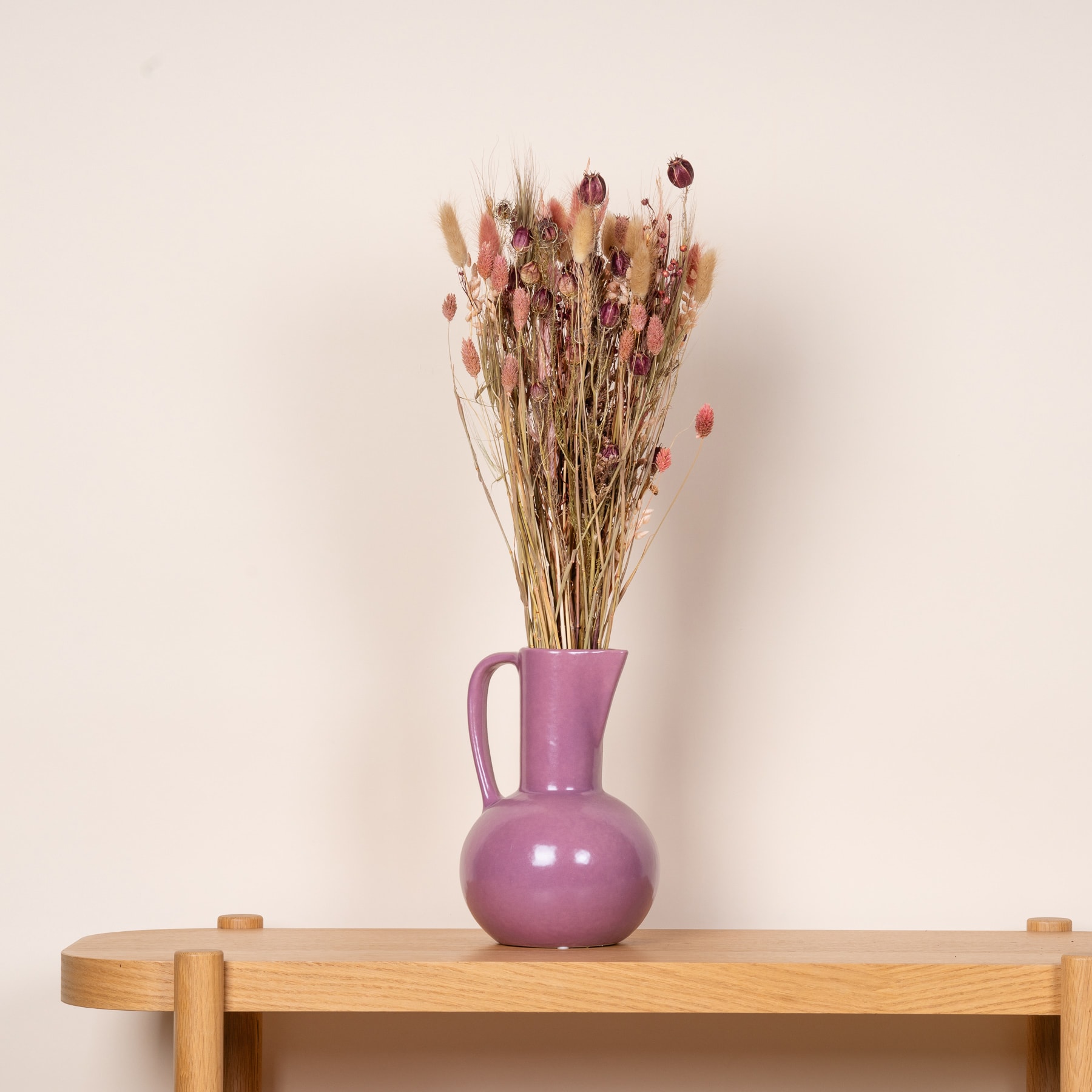 Ein farbenfroher Strauß getrockneter Blumen in einer lavendelfarbenen Keramikvase steht vor einem neutralen Hintergrund auf einem Holztisch.