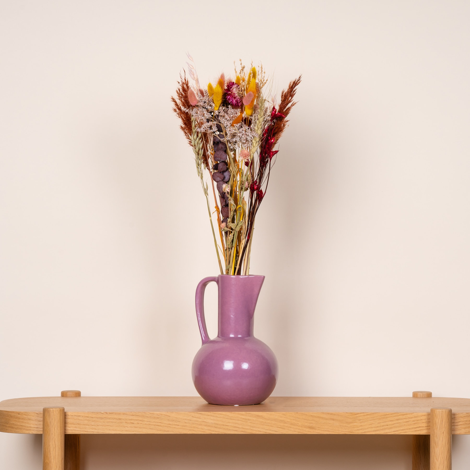Ein farbenfroher Strauß getrockneter Blumen in einer violetten Keramikvase steht auf einem Holztisch vor einem schlichten beigen Hintergrund.