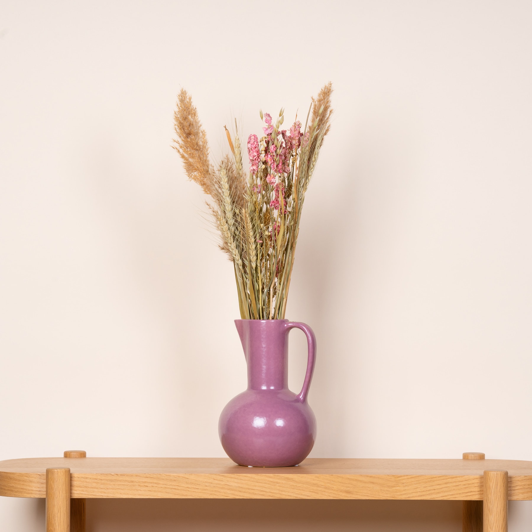 Ein farbenfroher Strauß getrockneter Blumen, darunter Pampasgras und rosa Blüten, wird in einer violetten Keramikvase auf einem Holzregal vor einem schlichten beigen Hintergrund präsentiert.