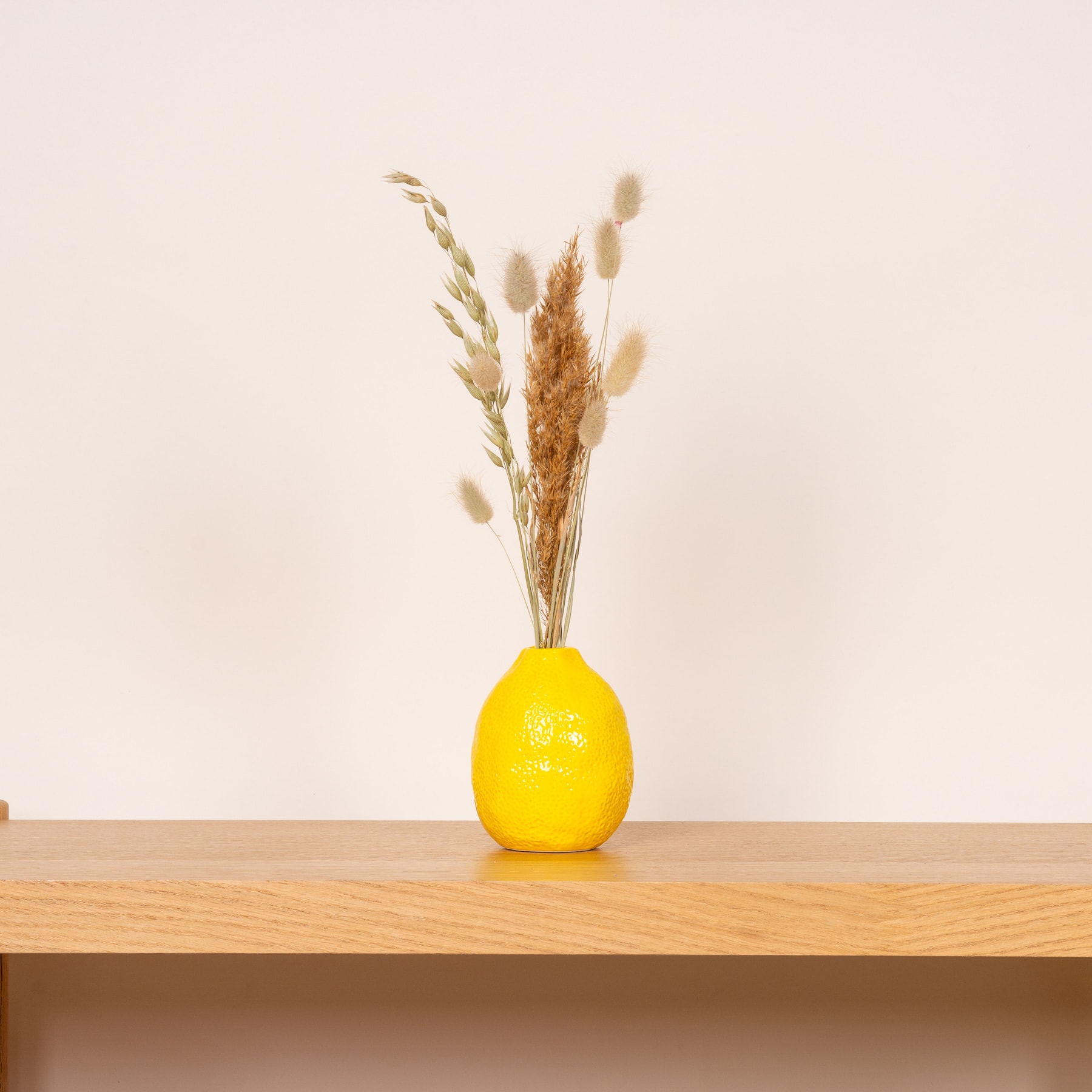 Eine gelbe Zitrone liegt auf einer Holzoberfläche, umgeben von getrockneten Blütenstängeln in einer minimalistischen Umgebung in neutralen Tönen.