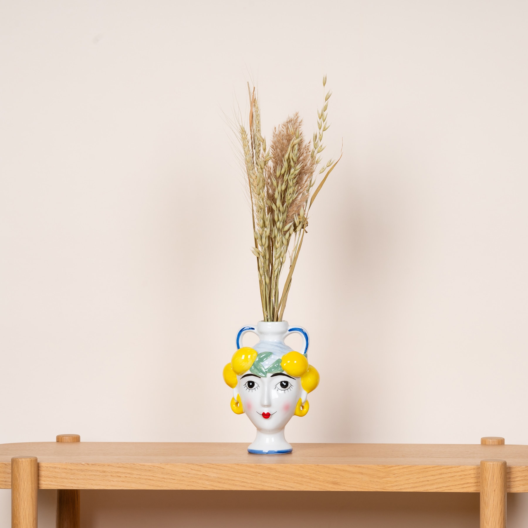 Eine Keramikvase mit einem skurrilen Gesichtsdesign enthält einen Strauß getrockneter Weizenhalme vor einem schlichten beigen Hintergrund.