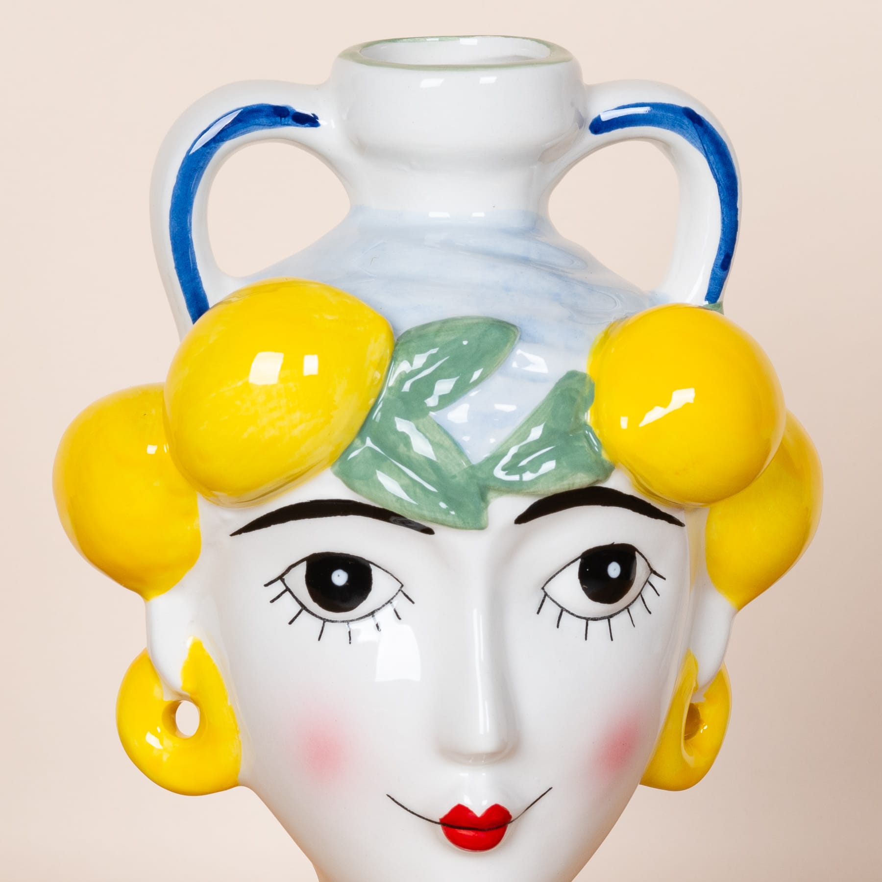 Eine Vase oder ein Gefäß aus Keramik mit einem Gesicht, das einer Frau ähnelt, mit großen gelben, zitronenähnlichen Formen an den Seiten und einem weißen Hintergrund.
